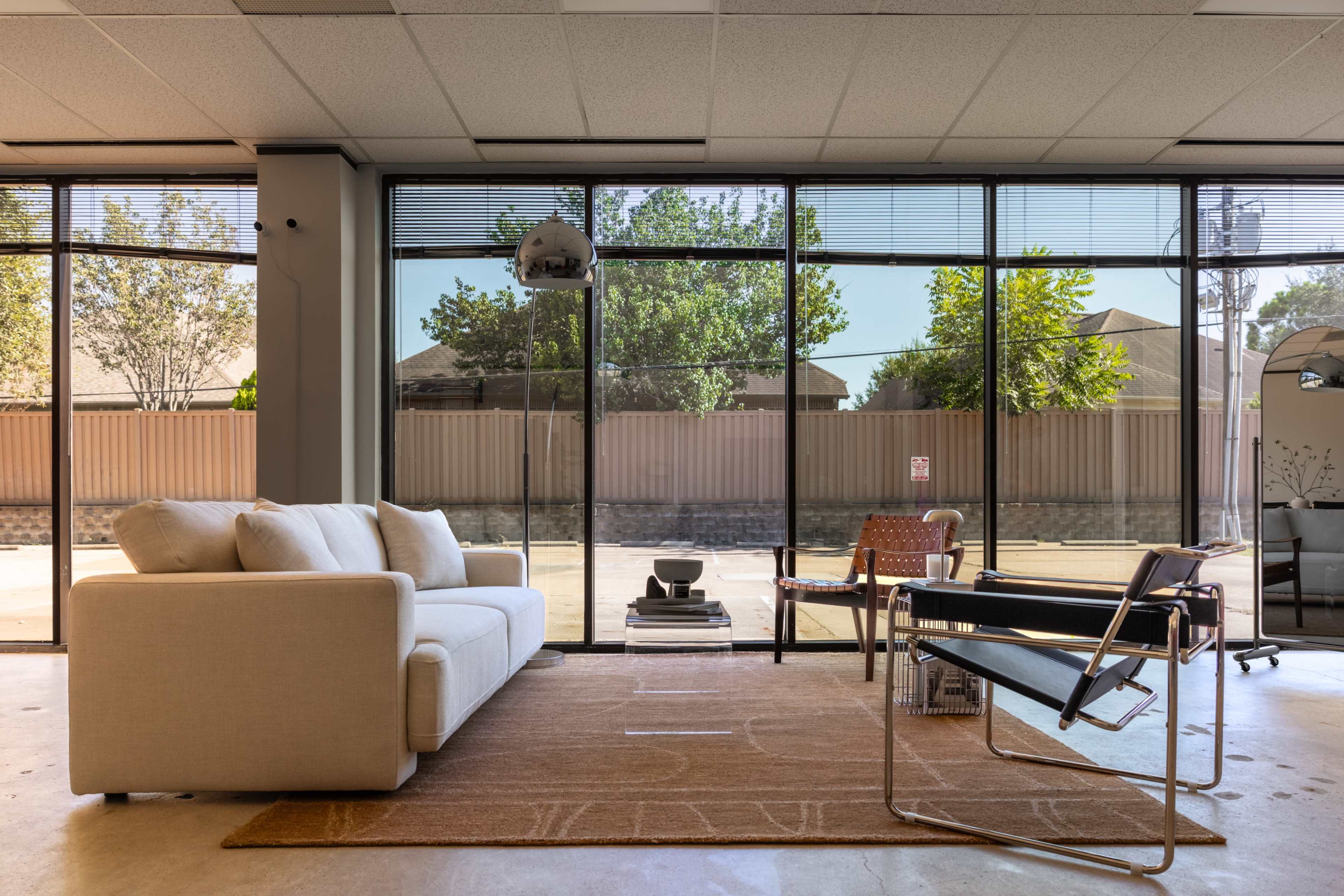 The image shows a modern living space with large windows, a beige sofa, a black chair, and a coffee table on a light rug.