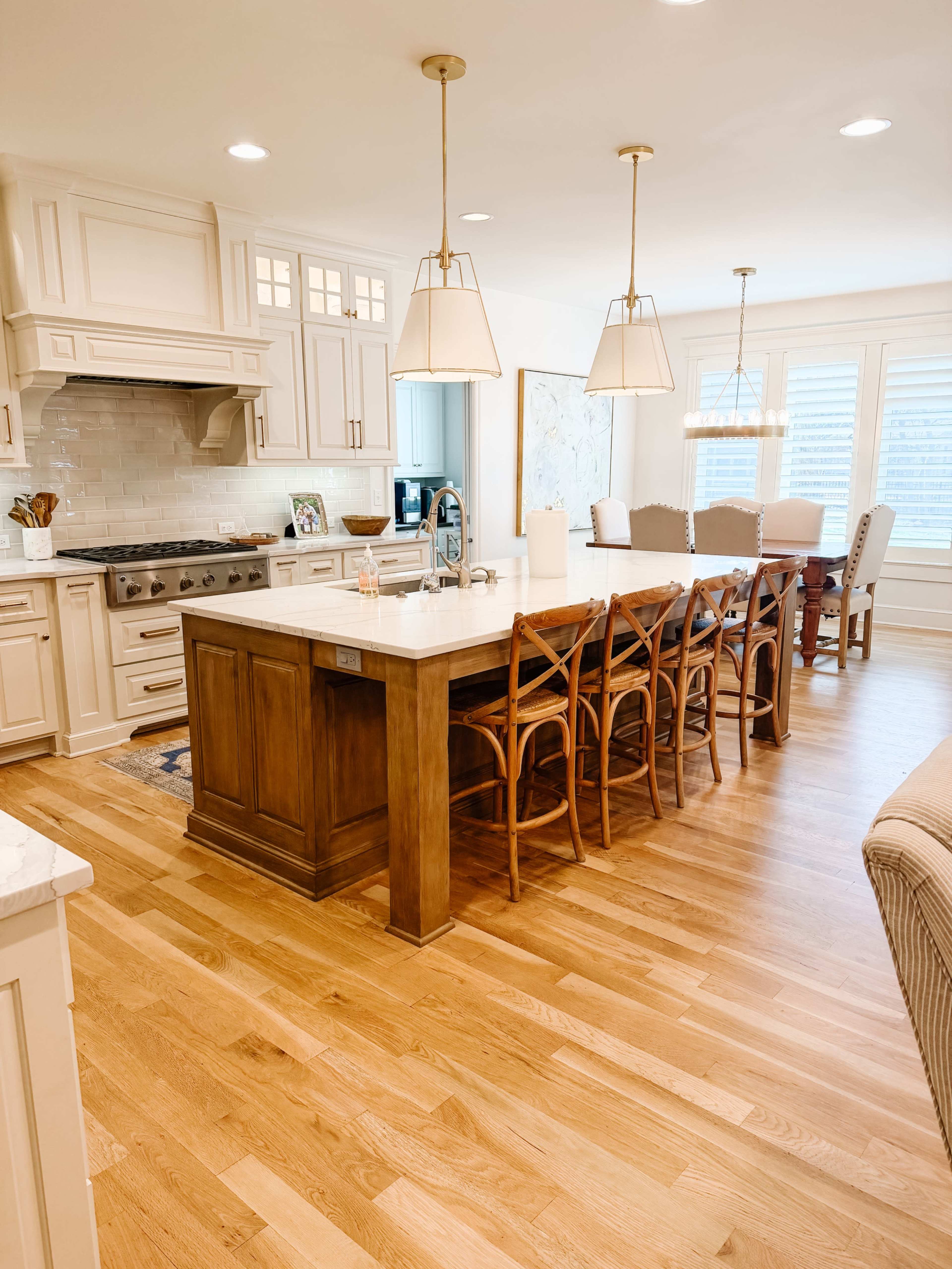 A modern kitchen features white cabinetry, a central island with bar stools, and an adjacent dining area with a large table under pendant lighting.