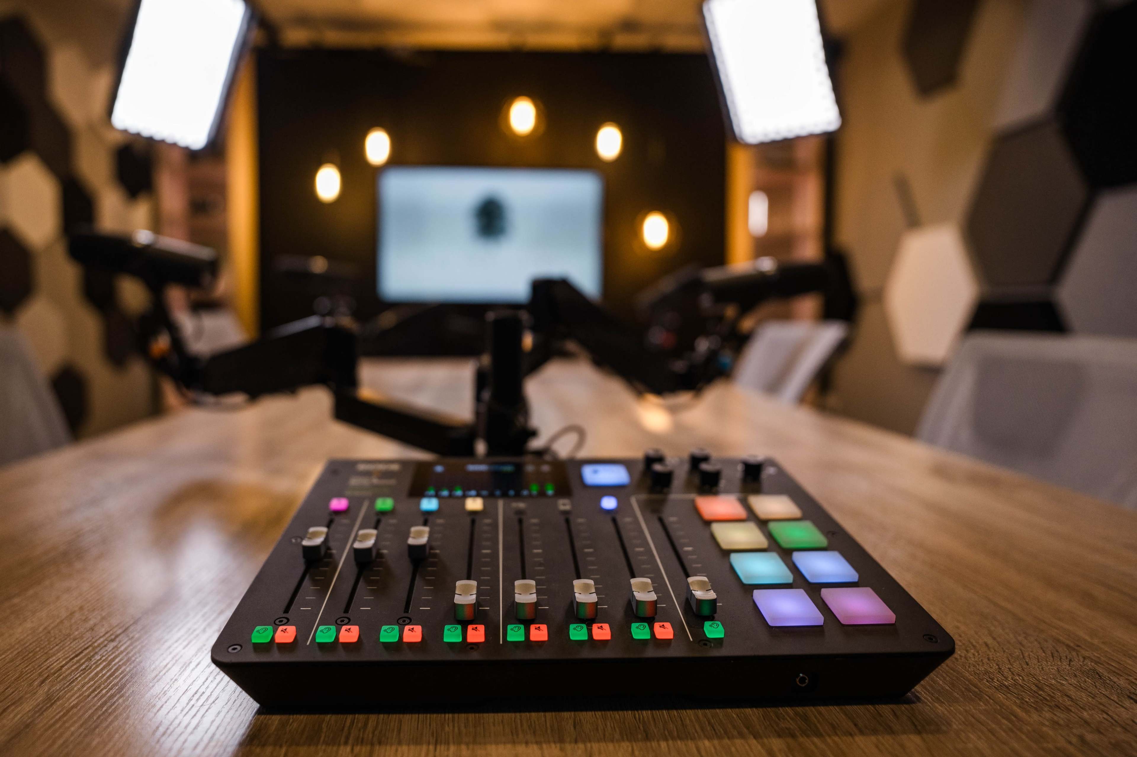 A close-up of a sound mixing console sits on a wooden table in a modern recording studio with overhead lights and a screen in the background.