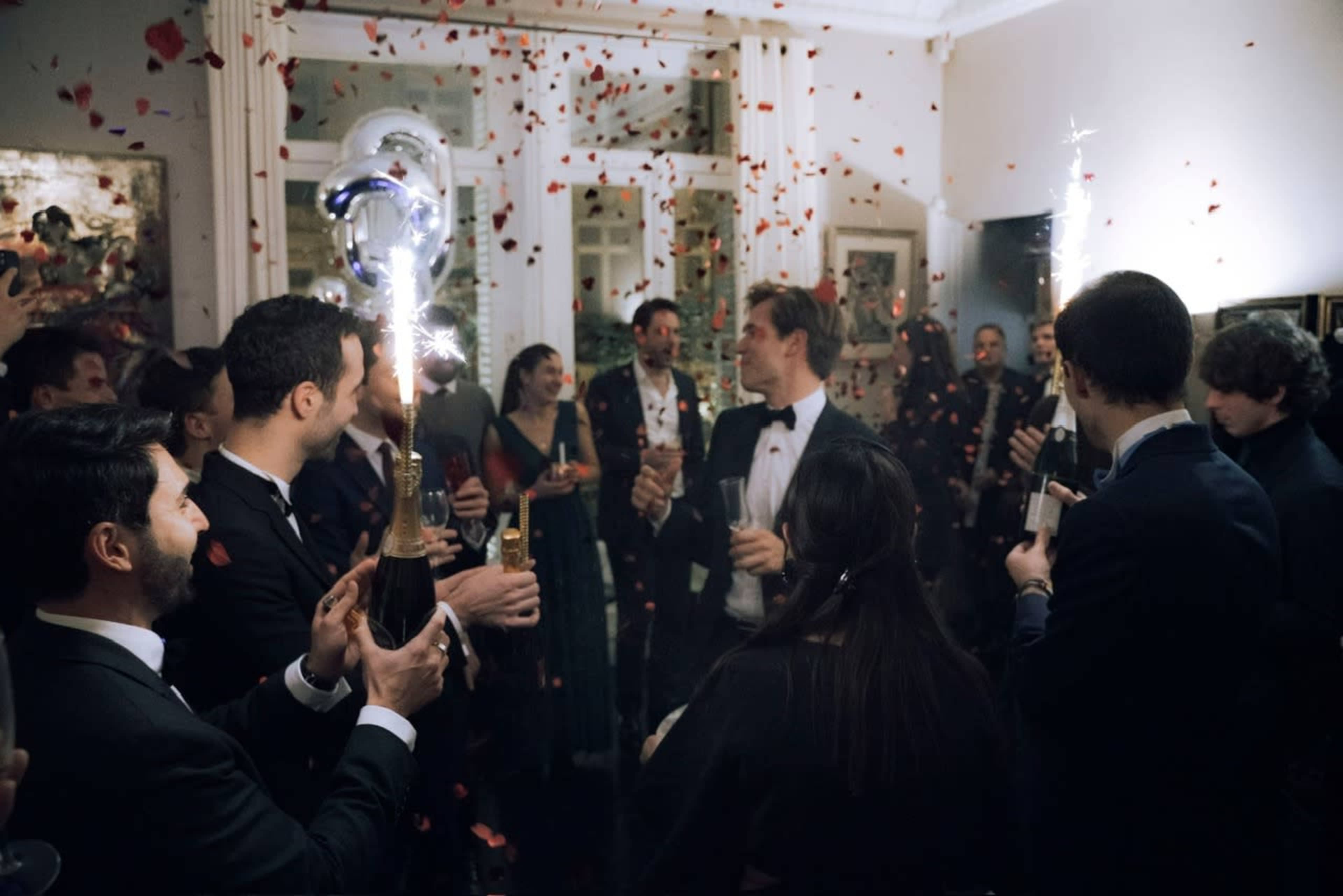 A group of elegantly dressed people celebrate at a party, holding champagne glasses and surrounded by confetti.