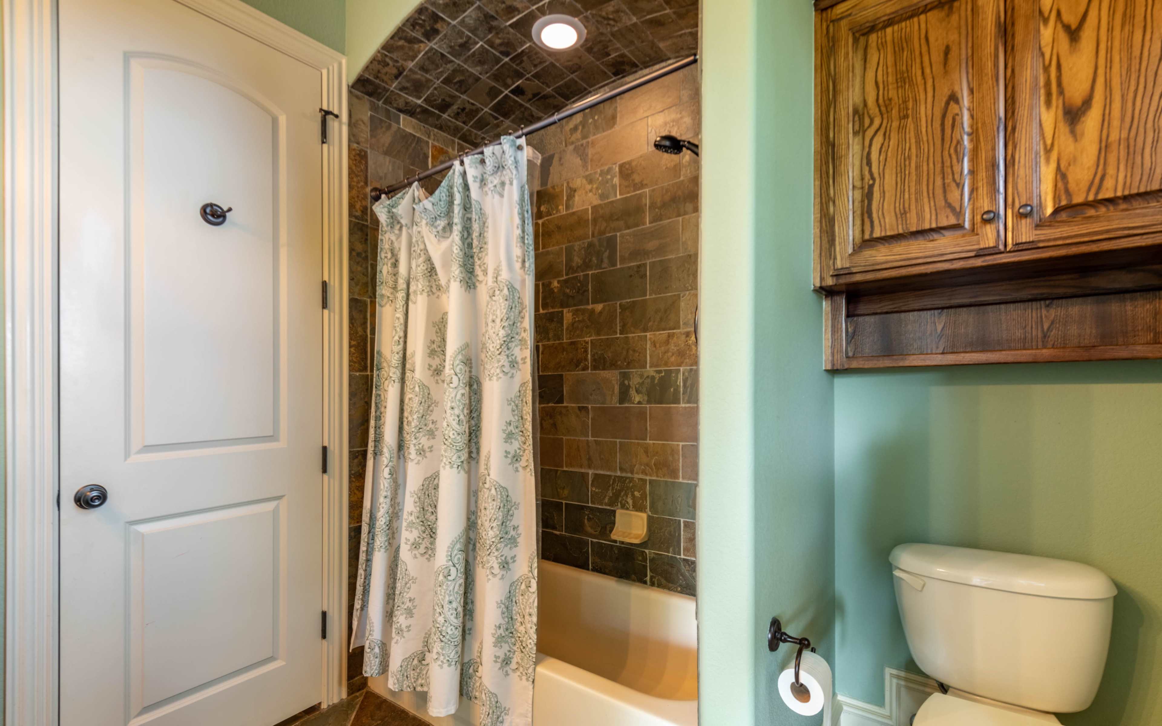 A bathroom featuring a shower with a curtain, a toilet, and a wooden cabinet, with tiled walls and a light green paint color.