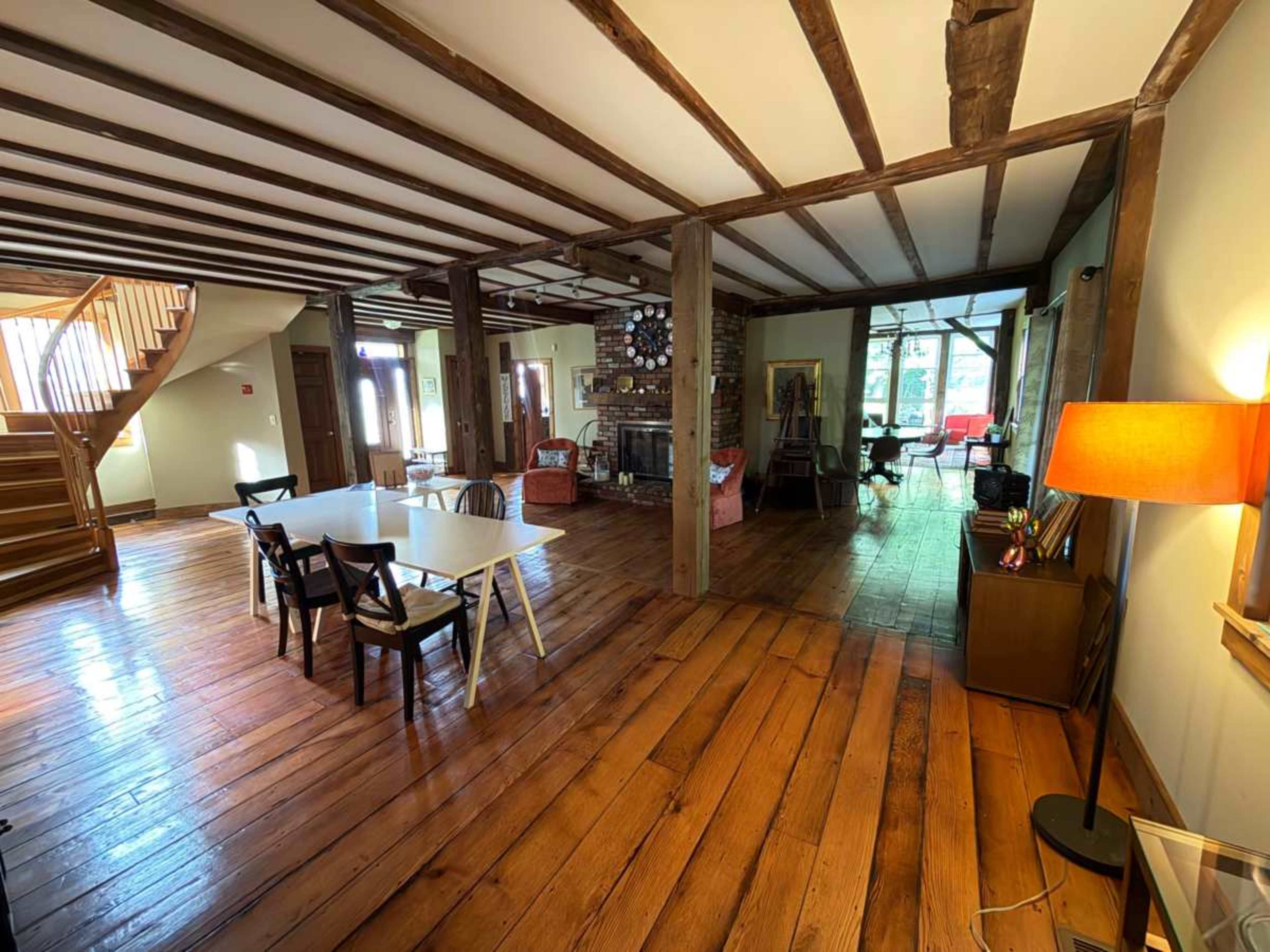 The image shows a spacious, well-lit interior with wooden beams, a cozy seating area, and a dining table set for multiple people.
