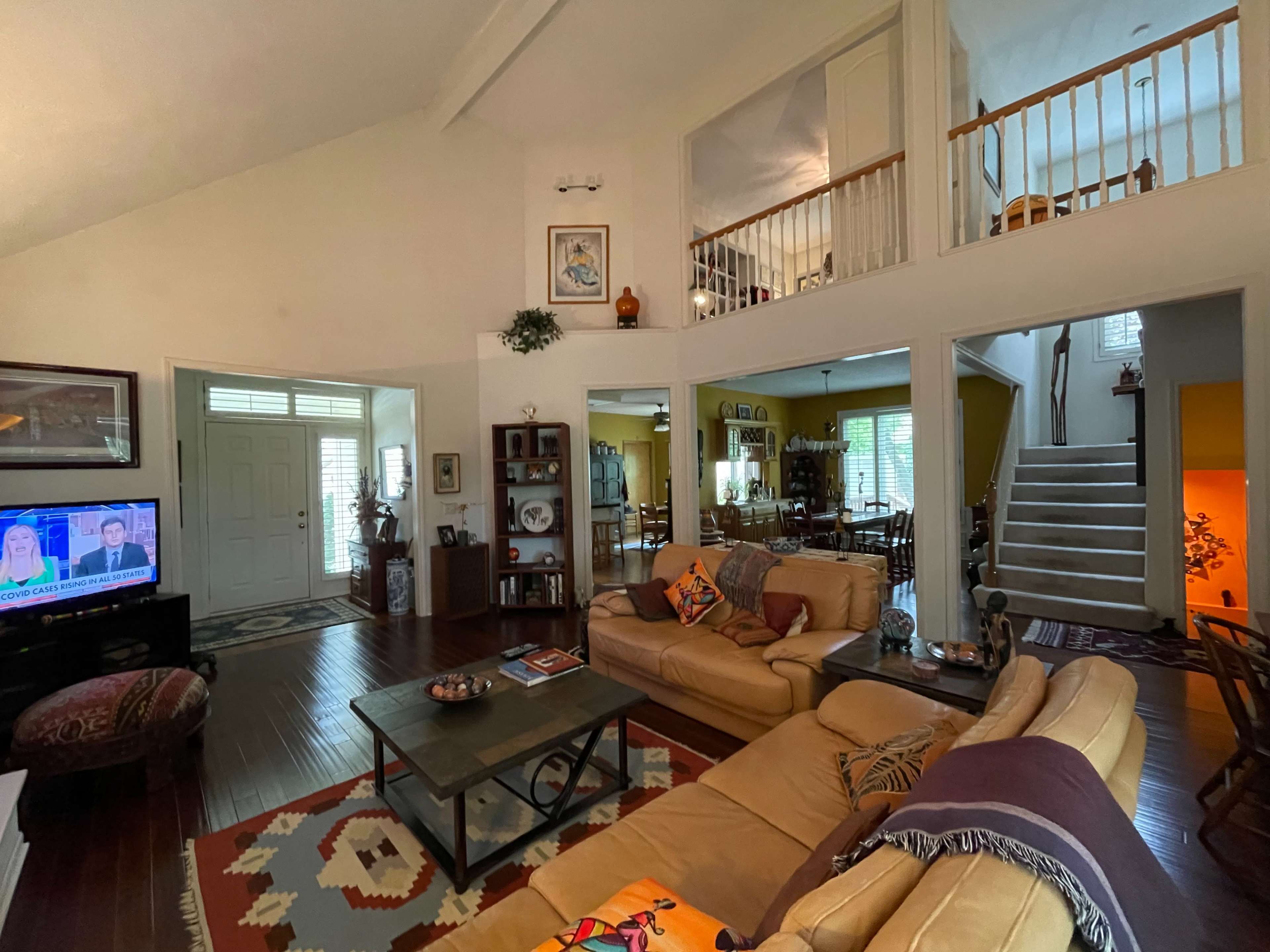 The living room features a beige sofa set, a coffee table, and a large television, creating an open space that connects to a dining area and a staircase.