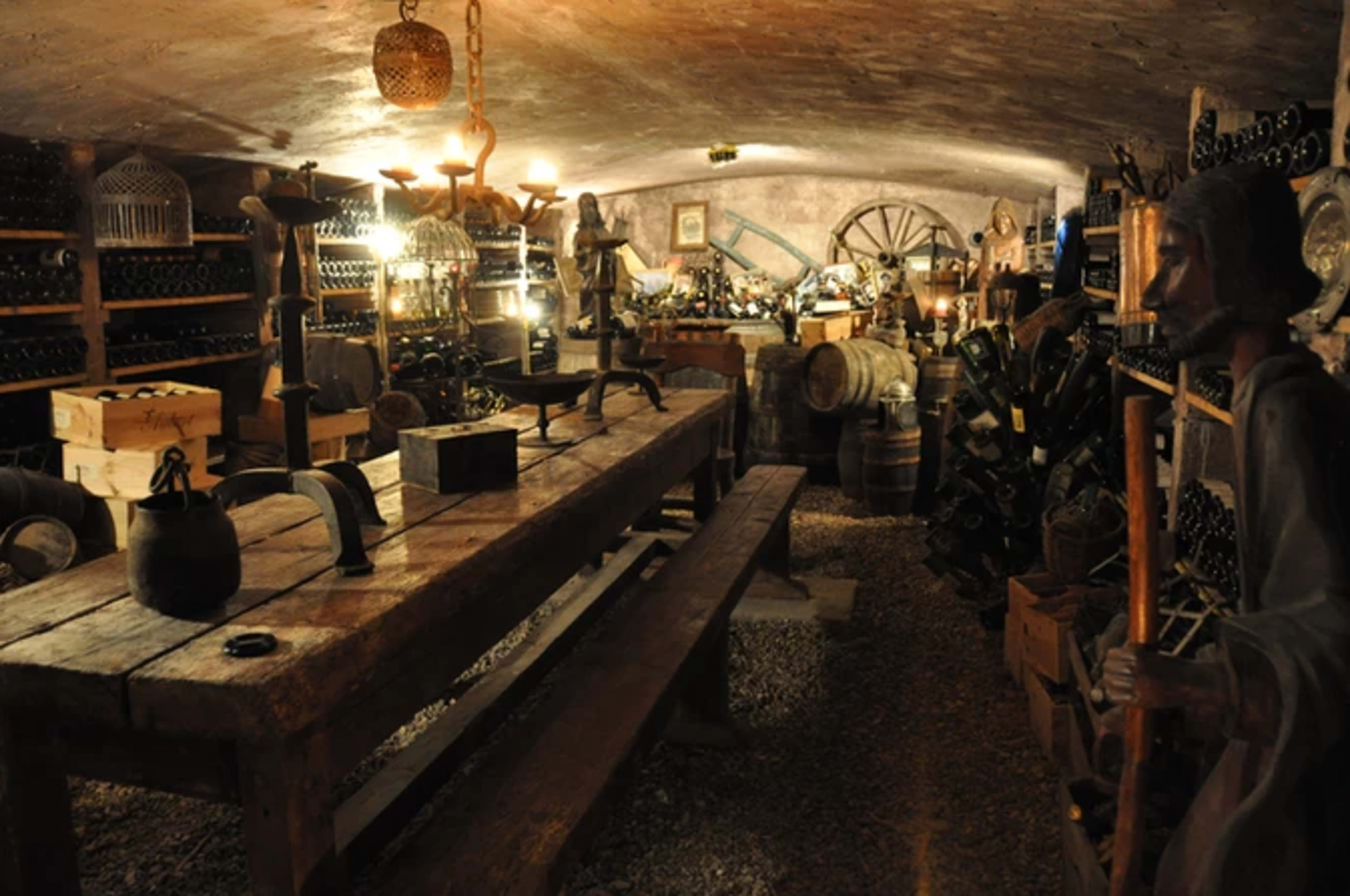 A dimly lit cellar filled with wooden shelves stacked with wine bottles, antique tools, and wooden barrels, with a large table in the center.