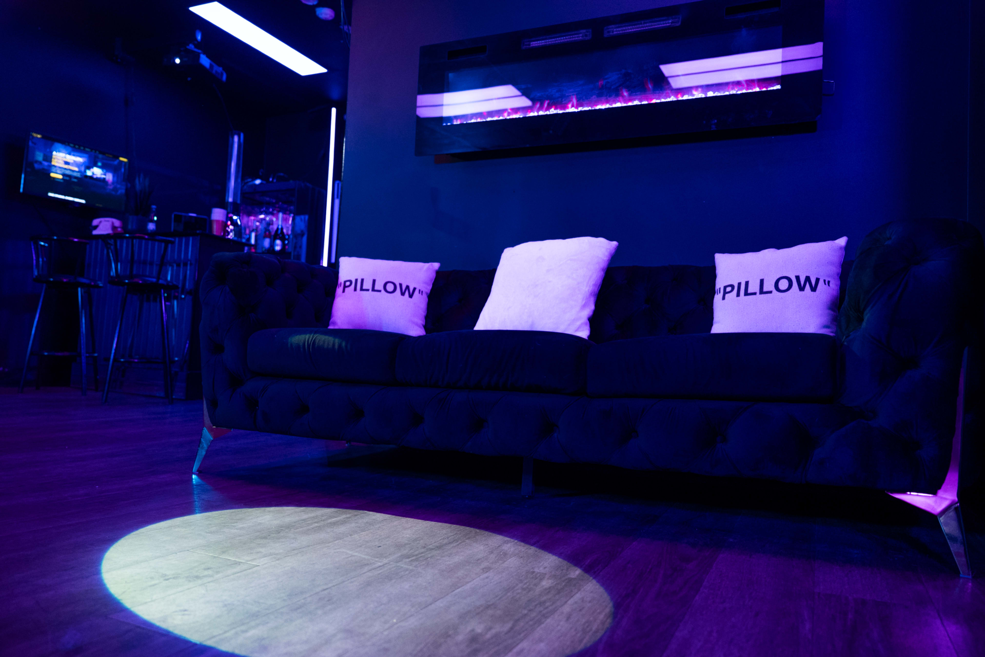 A black tufted couch with two decorative pillows sits under a purple-lit wall featuring a modern fireplace in a dimly lit room.