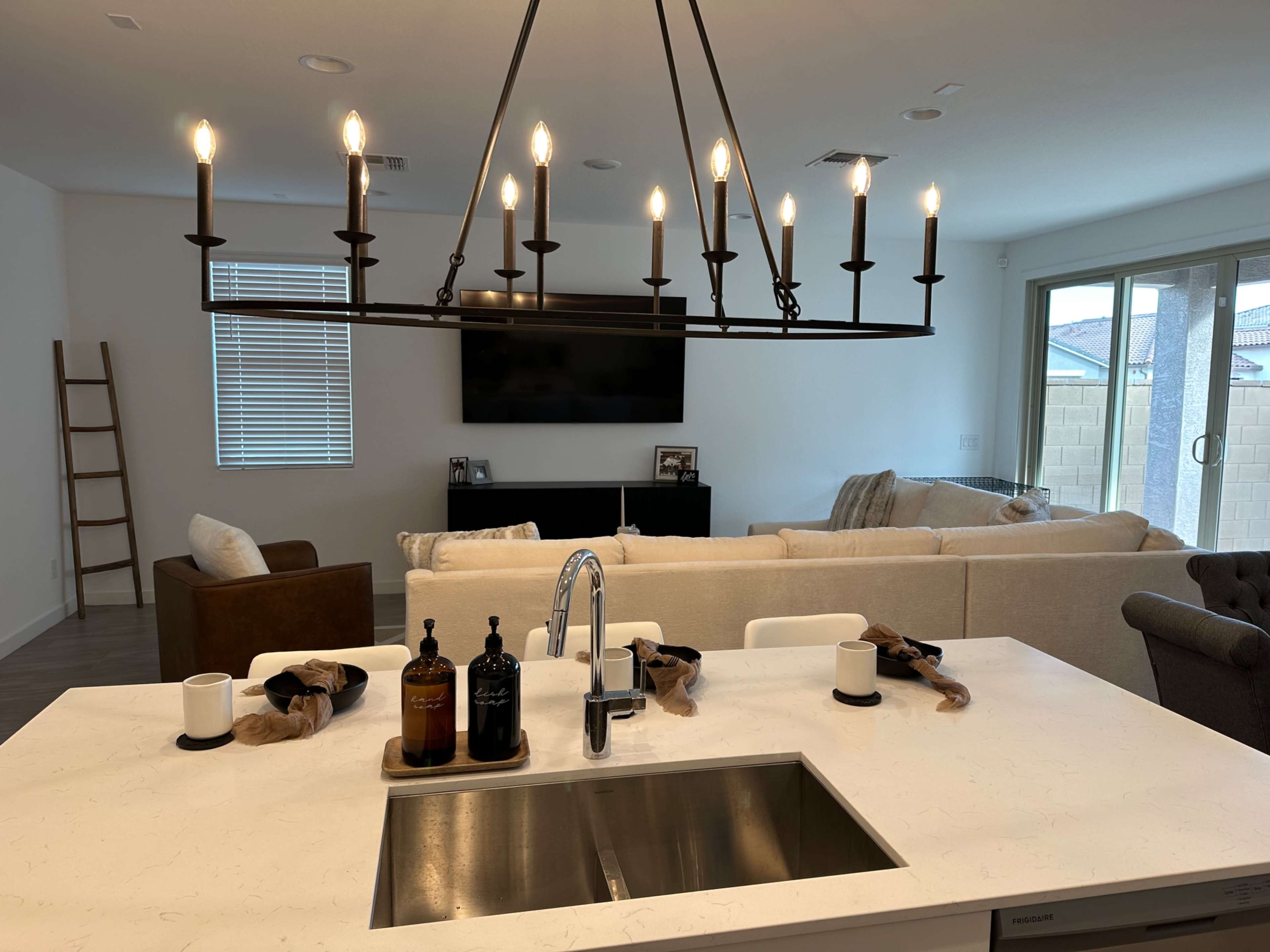 A modern kitchen features a large island with a sink, accompanied by decorative bottles, while a spacious living room with a sofa and a media console is visible in the background.