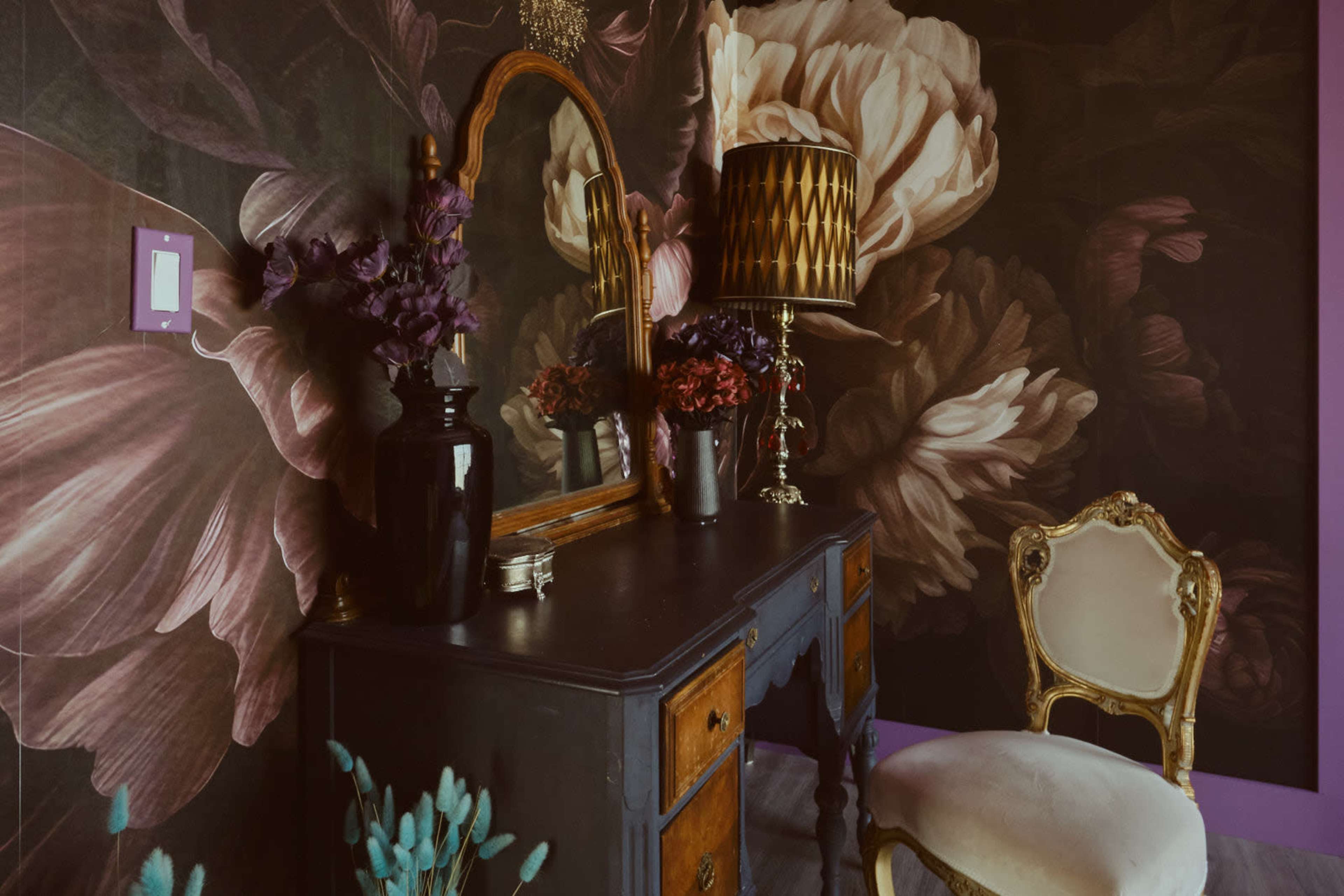 The image shows a darkly decorated room featuring a vintage dresser with a mirror, a lamp, and decorative flowers against a floral wallpaper backdrop.