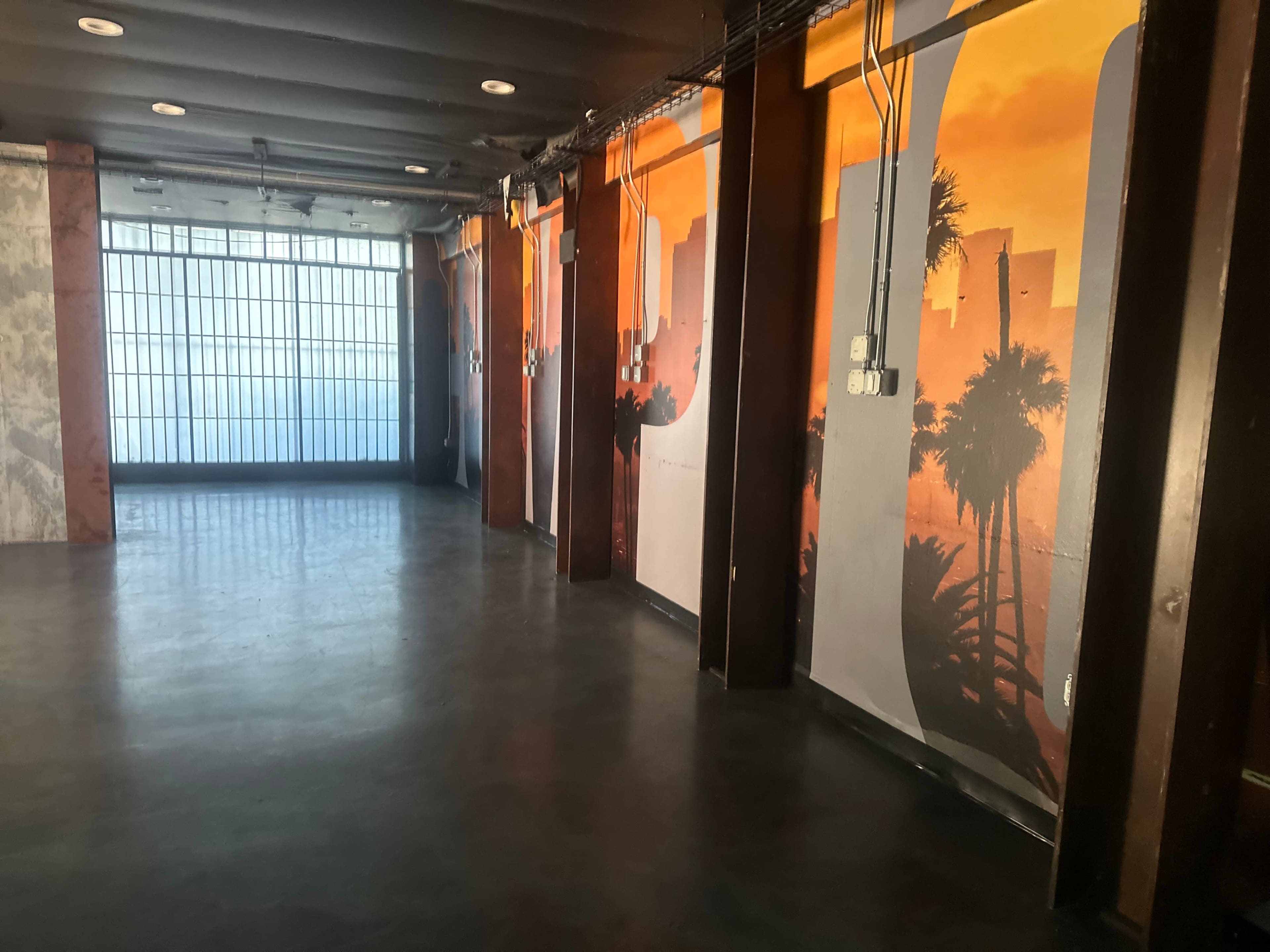 The image shows an empty interior space with large wall murals featuring a sunset cityscape and palm trees.