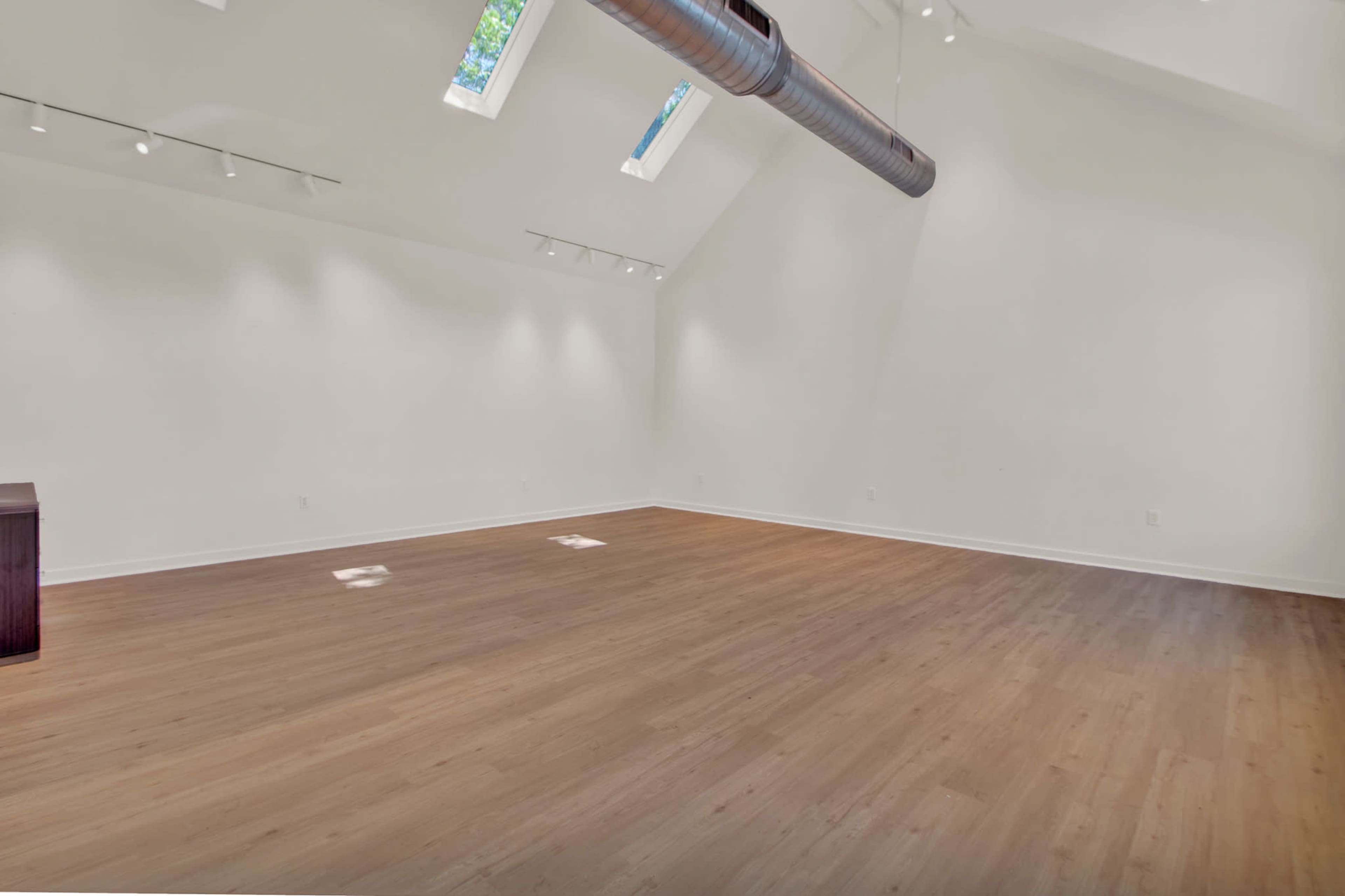 The image shows a spacious, minimally furnished interior with wooden flooring and large windows allowing natural light to enter.