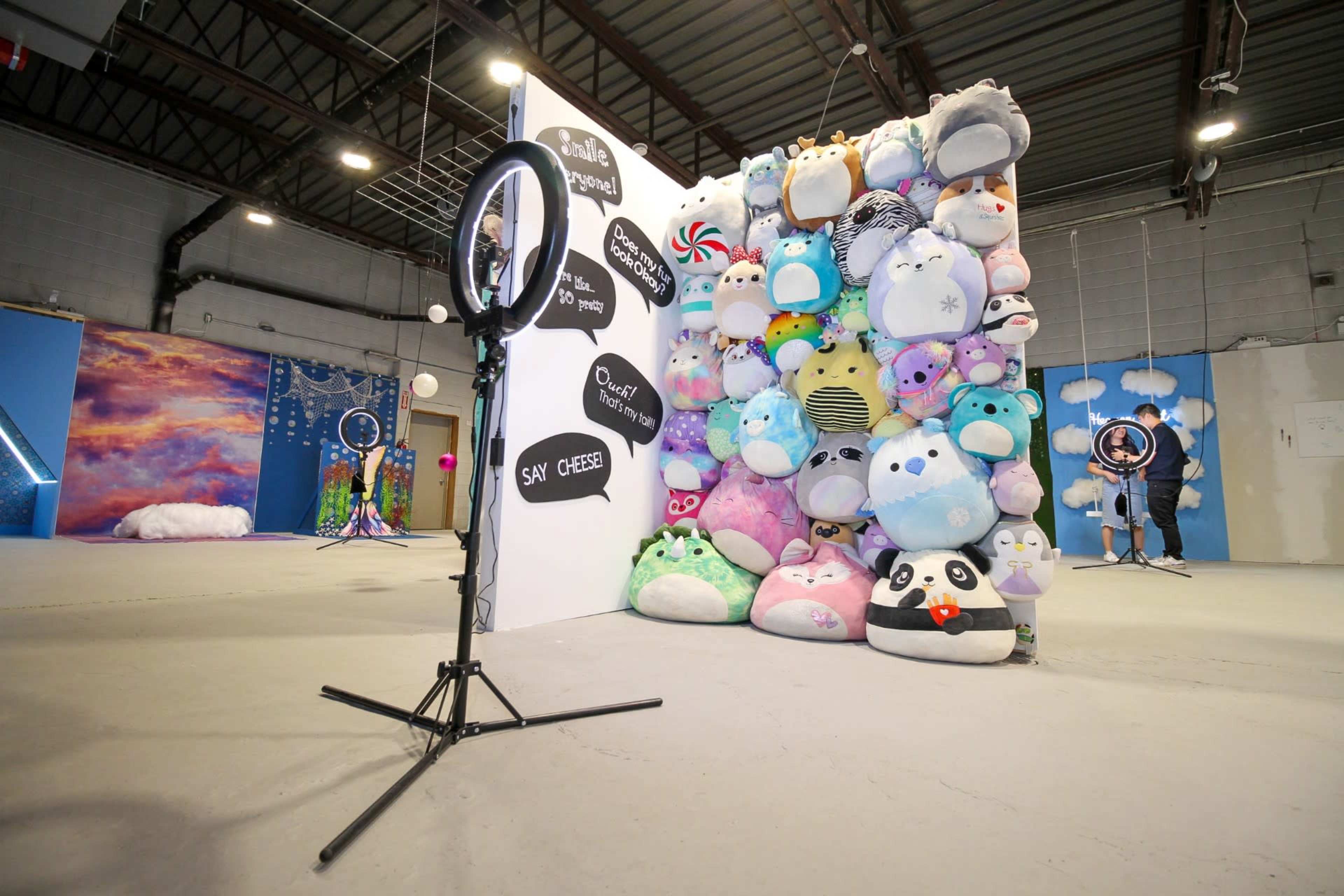 The scene features a colorful wall of plush stuffed animals alongside a ring light set up for photography.