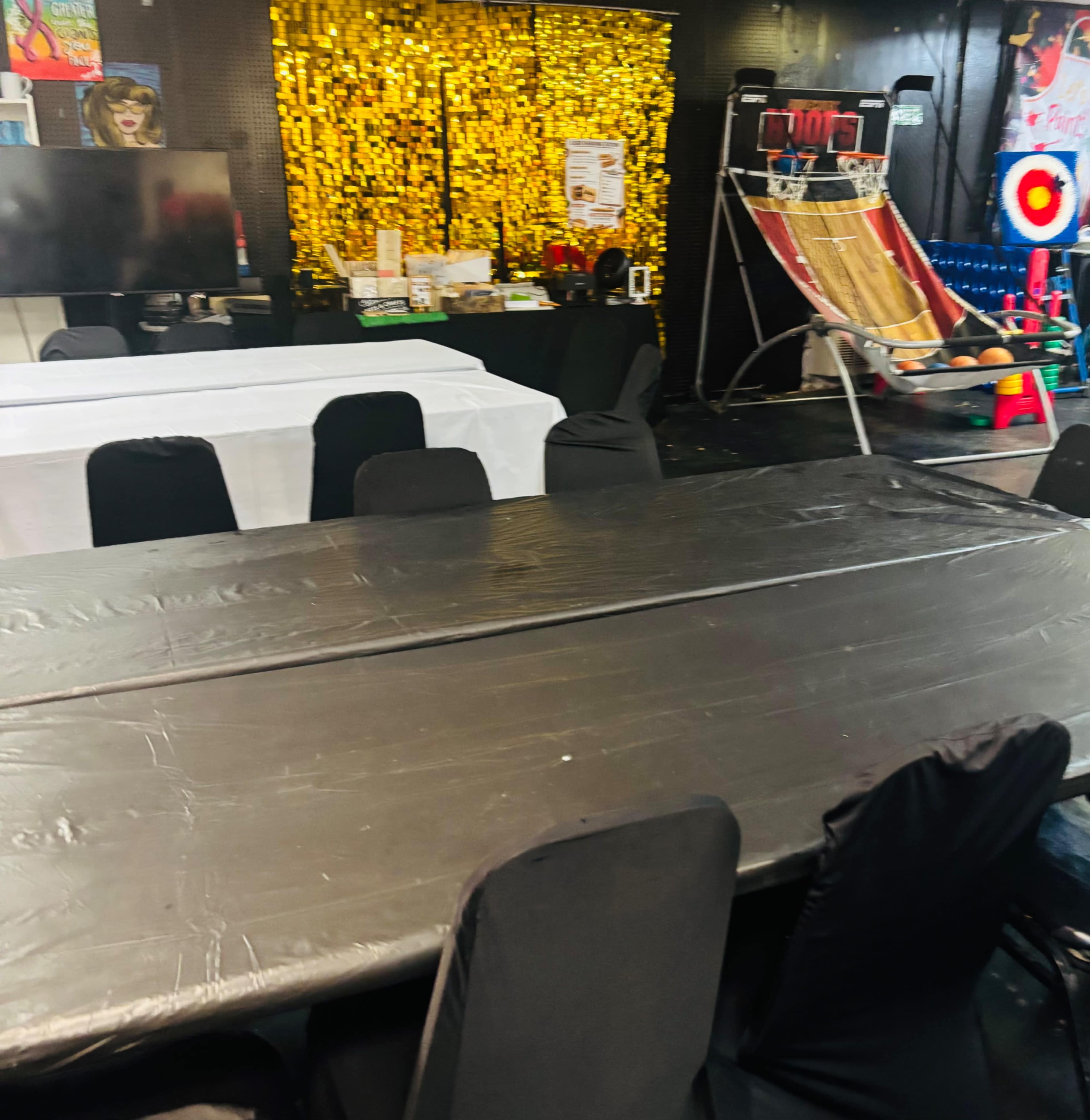 The image shows a room set up for an event, featuring a large black table covered with a plastic tablecloth, surrounded by black chairs, and a variety of entertainment equipment in the background, including a basketball shooting arcade game and a colorful backdrop.