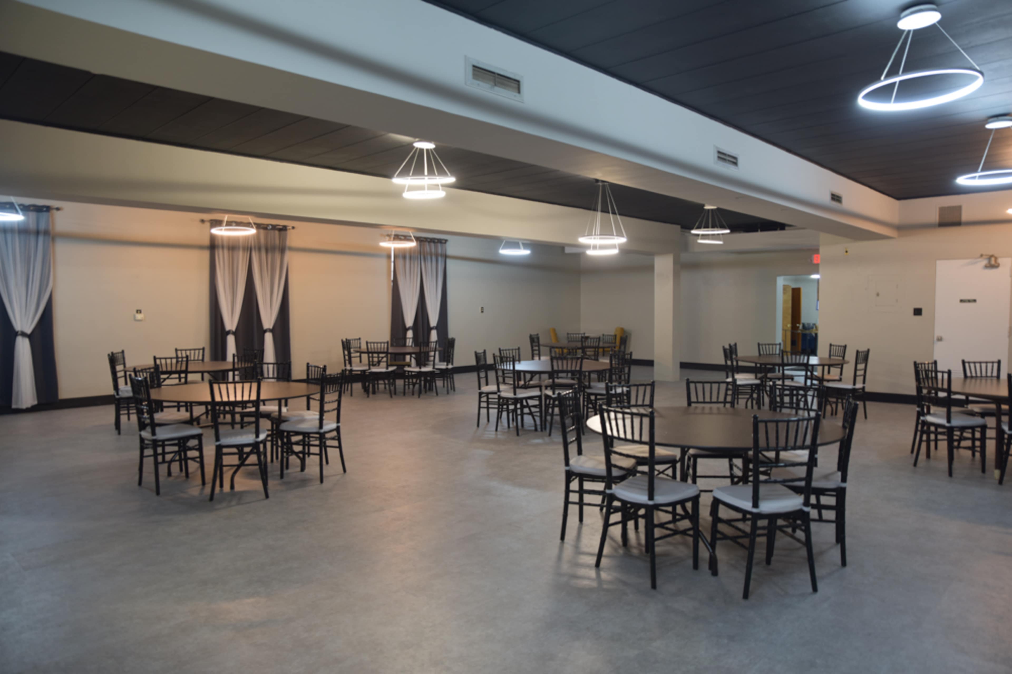 A spacious, empty dining hall with several round tables and black chairs arranged neatly under modern pendant lights.