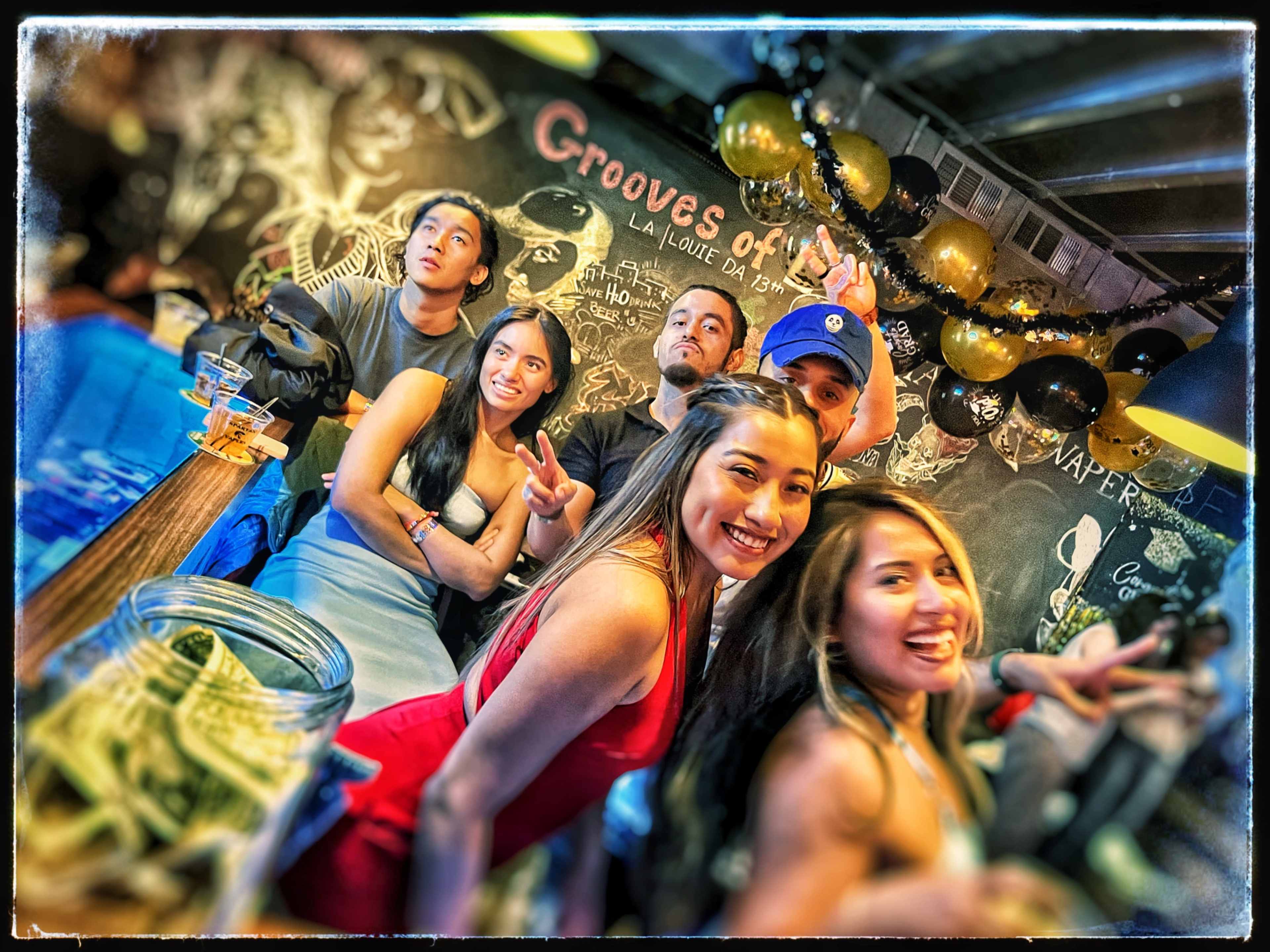 A group of six people poses playfully at a bar decorated with balloons and art on the black wall behind them.