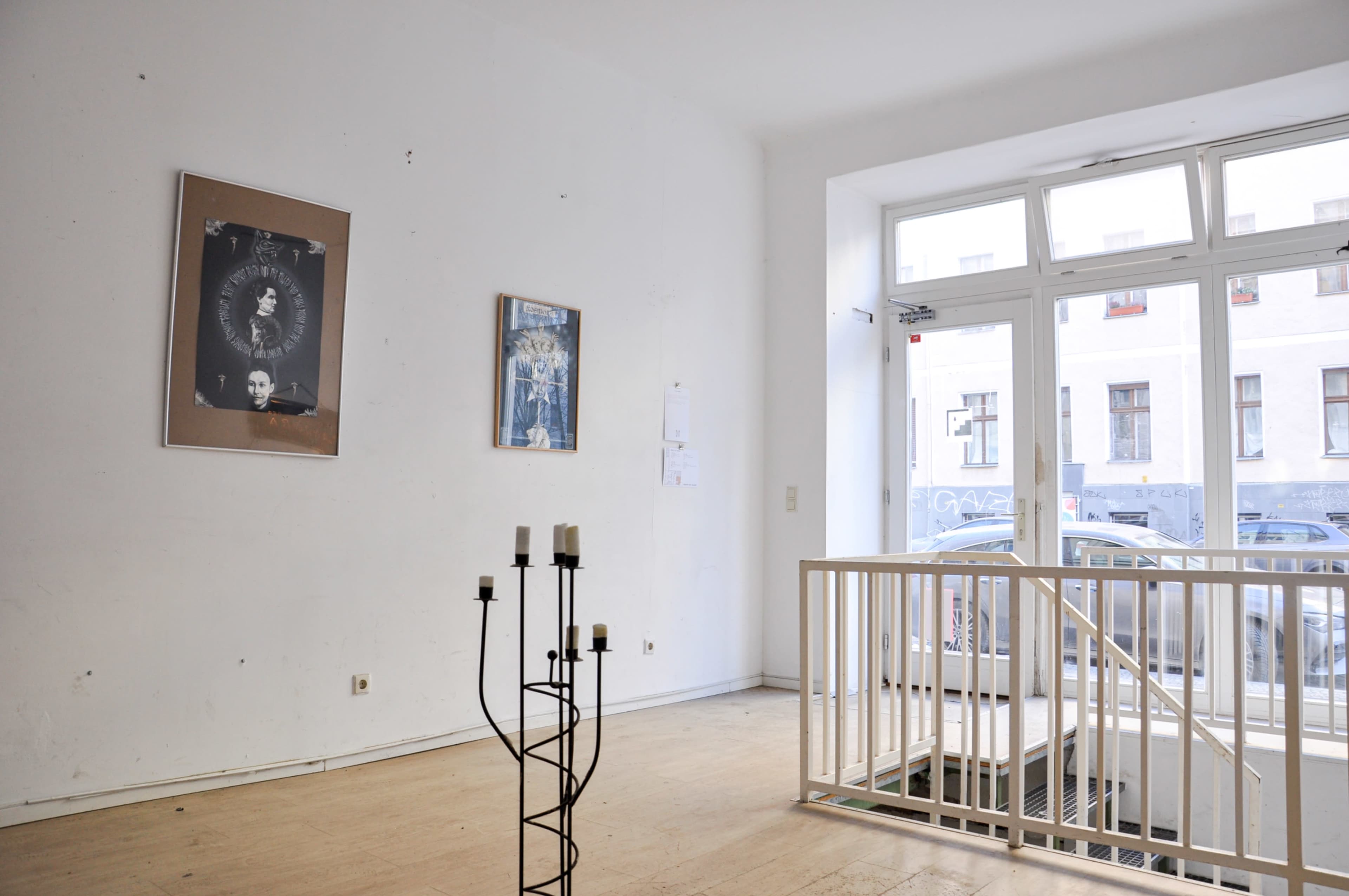 The image shows a sparse gallery space with two framed pictures on the white walls, a metal railing, and a doorway leading to an exterior street view.