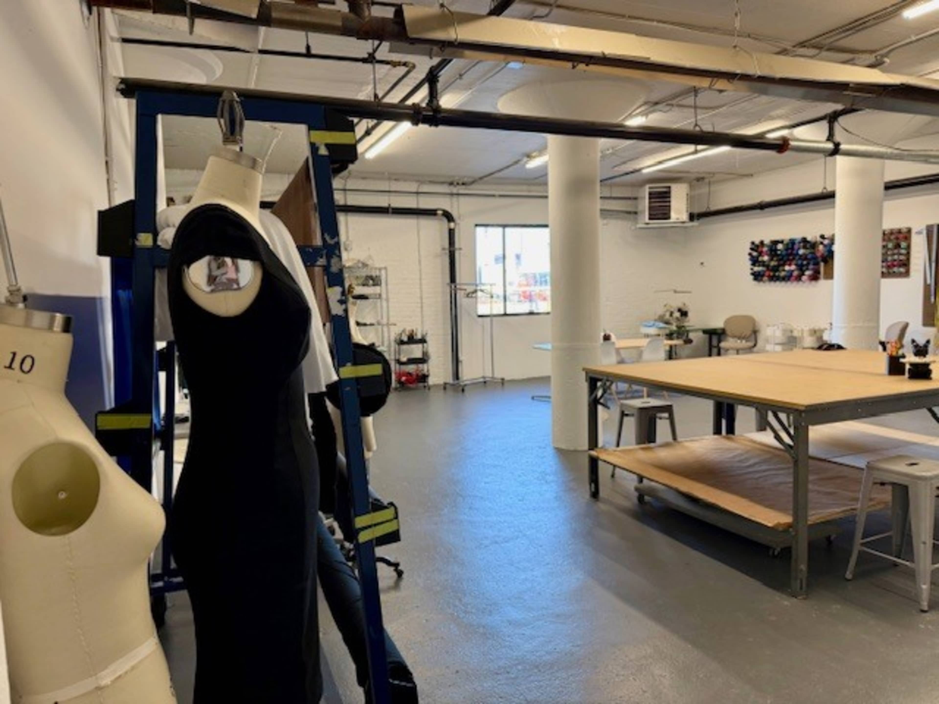 The image shows a spacious sewing studio with dress forms, tables, and sewing equipment positioned around the room.