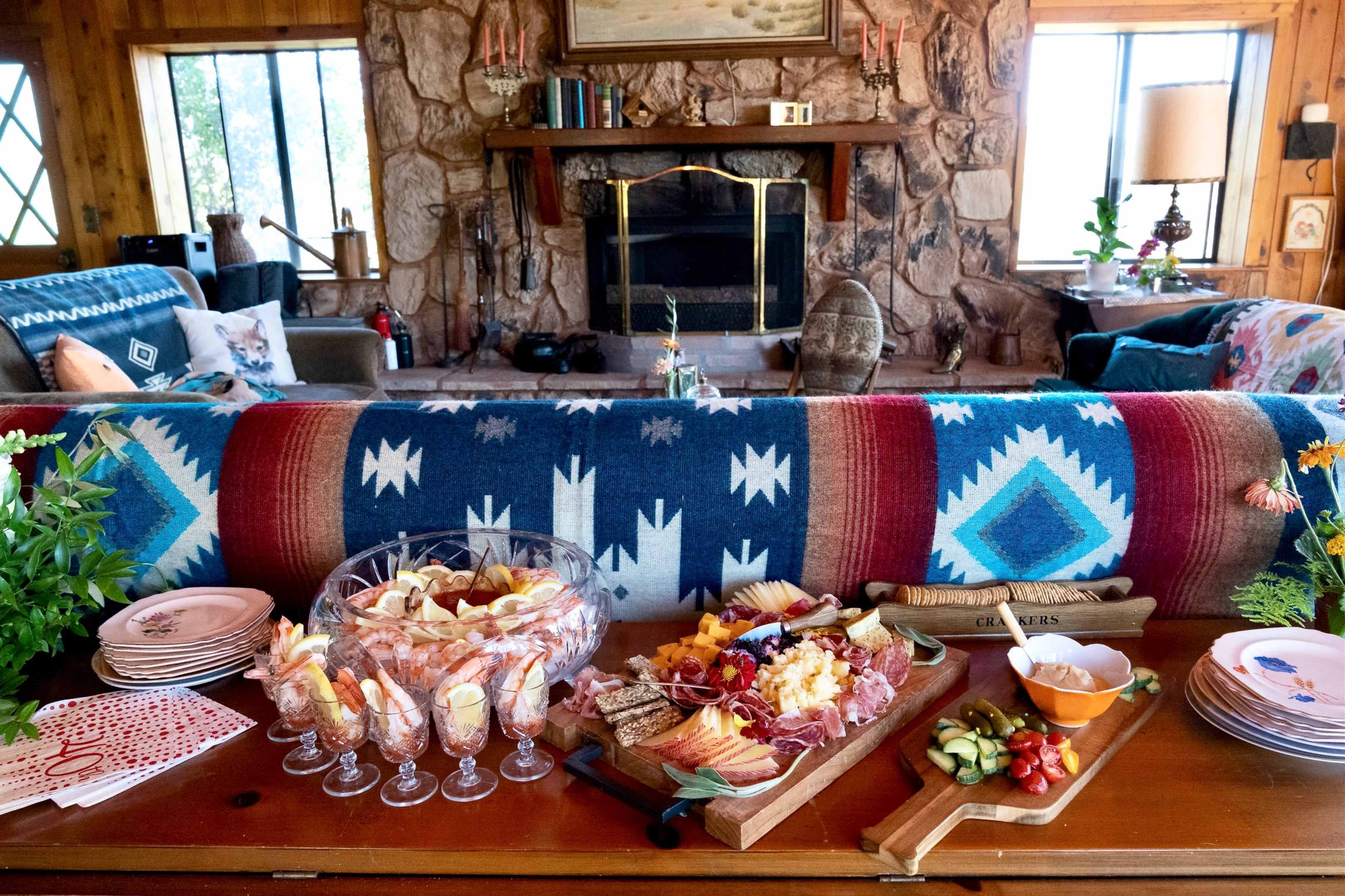 A rustic living room features a wooden table set with an assortment of appetizers, including a charcuterie board, fruit, and decorative glasses, alongside a cozy stone fireplace.