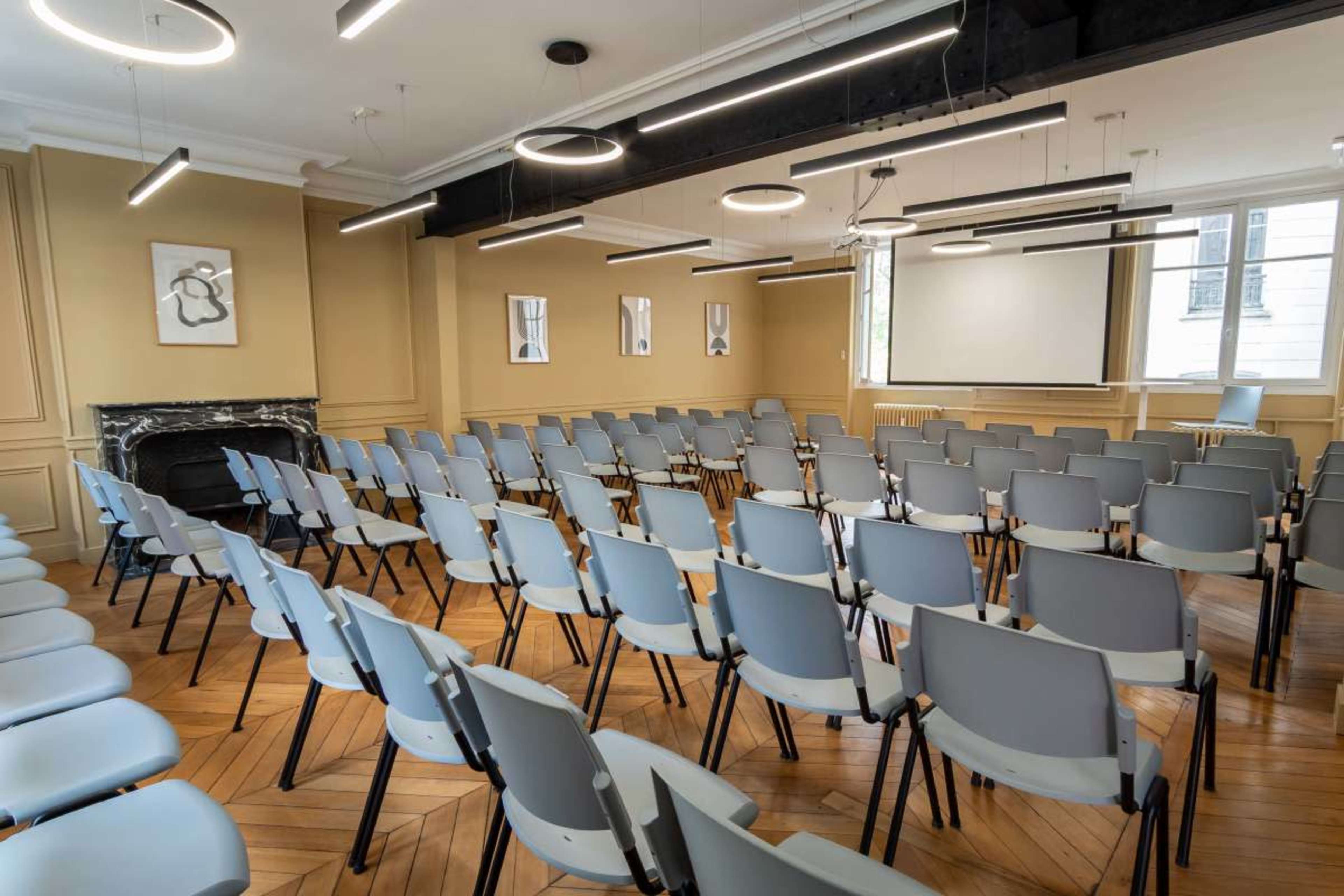 The image shows a conference room arranged with neatly positioned gray chairs facing a screen at the front.