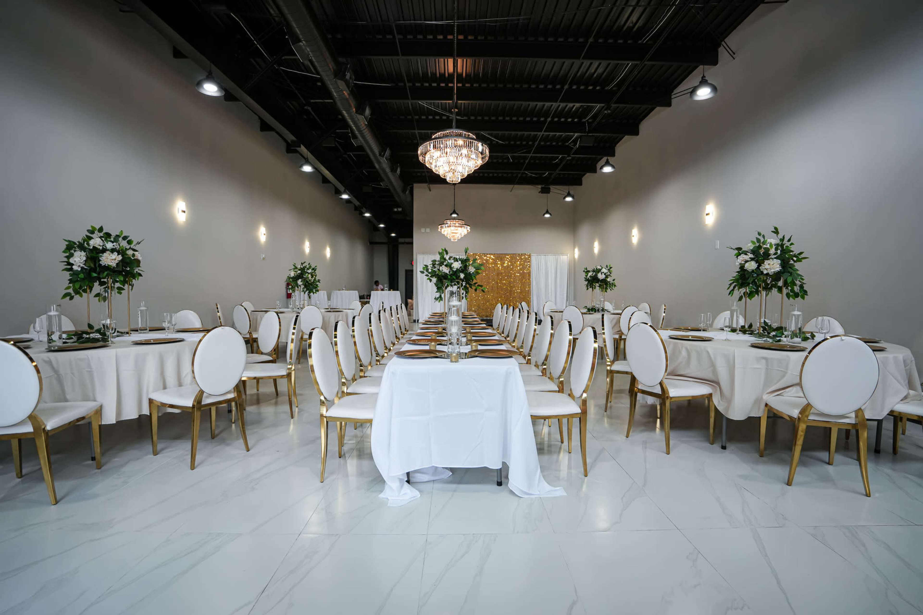 A spacious banquet hall is set up with round tables decorated with white tablecloths and gold-accented chairs, featuring centerpieces of greenery and crystal chandeliers overhead.