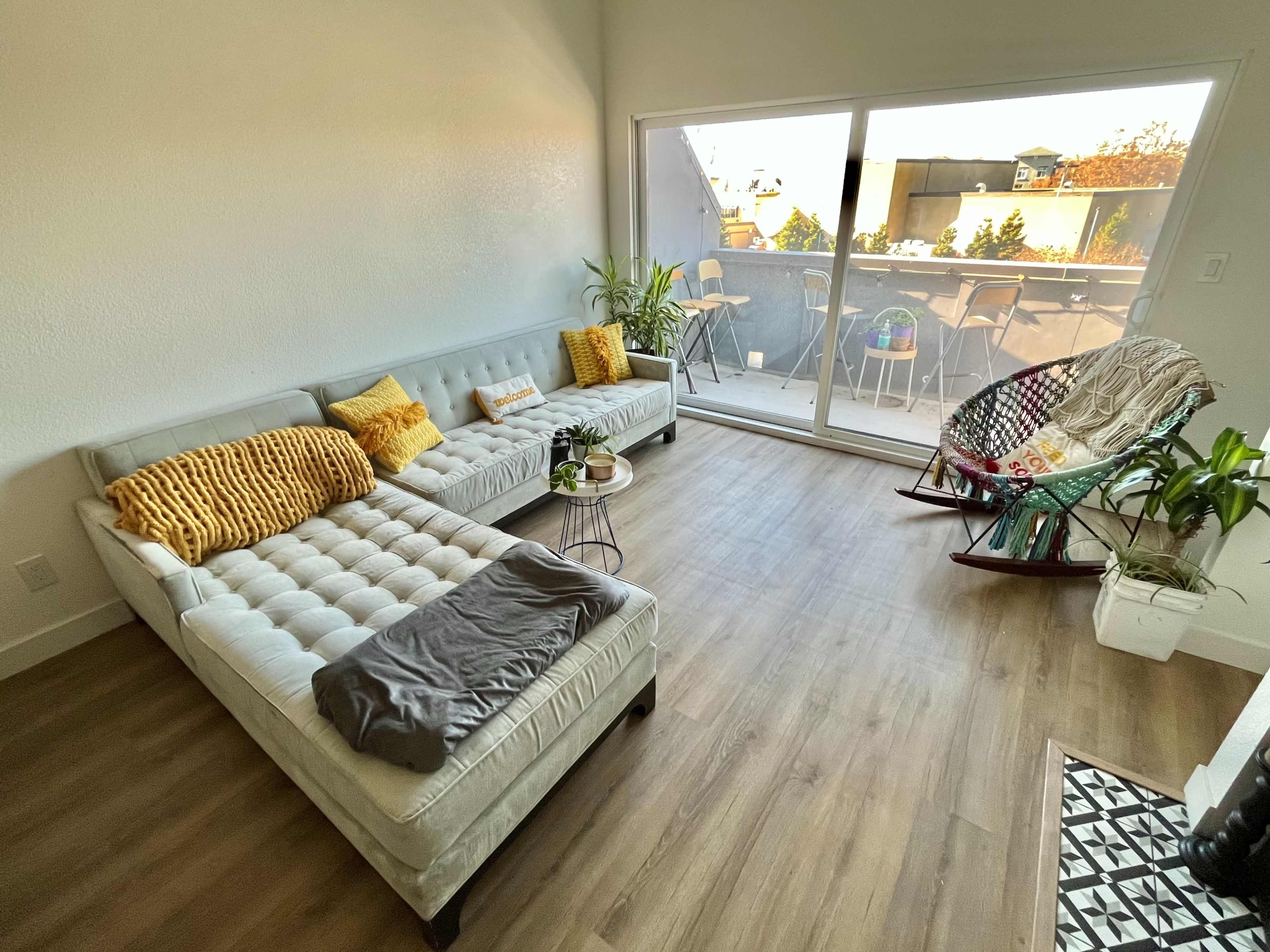 The room features a light-filled space with a gray sectional sofa, a rocking chair, a small table, and plants, leading to a balcony with outdoor seating.