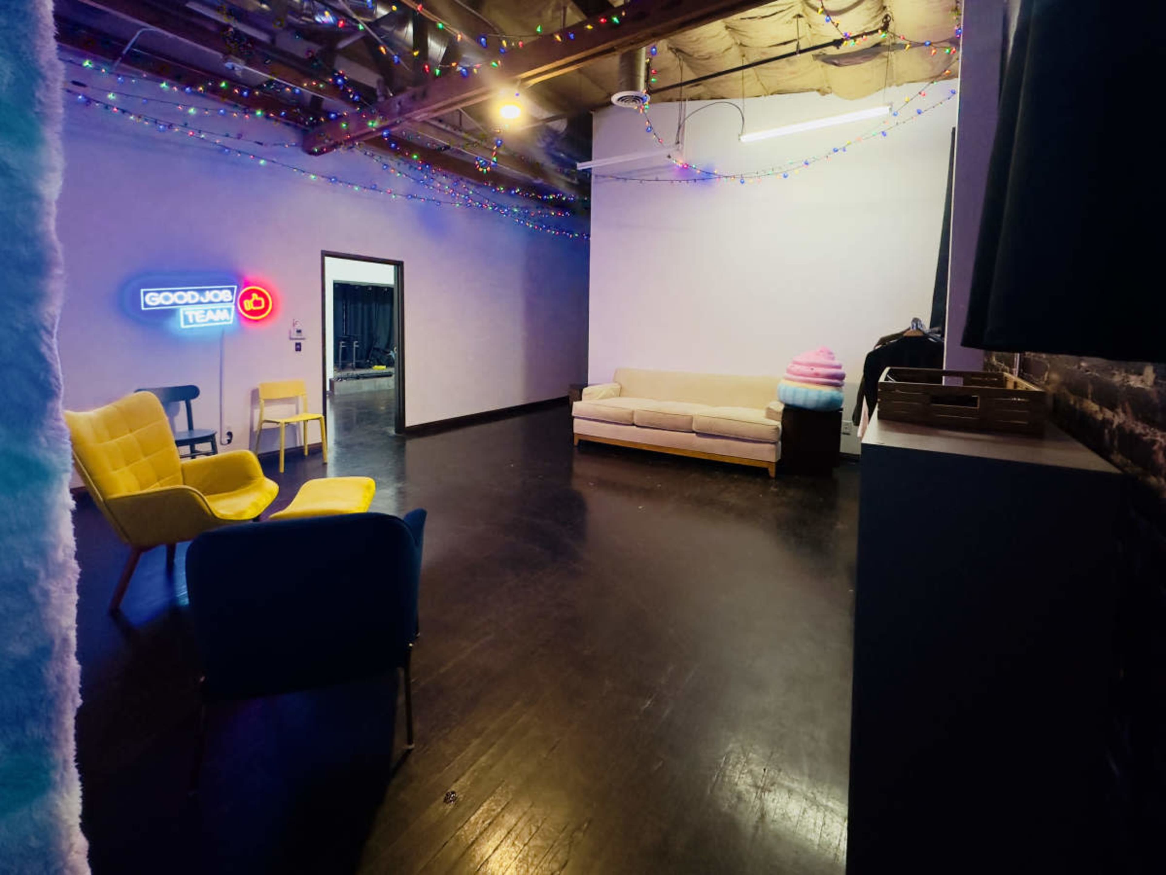 A spacious room with colorful lighting, featuring a beige sofa, two brightly colored armchairs, and a cotton candy machine in the corner.