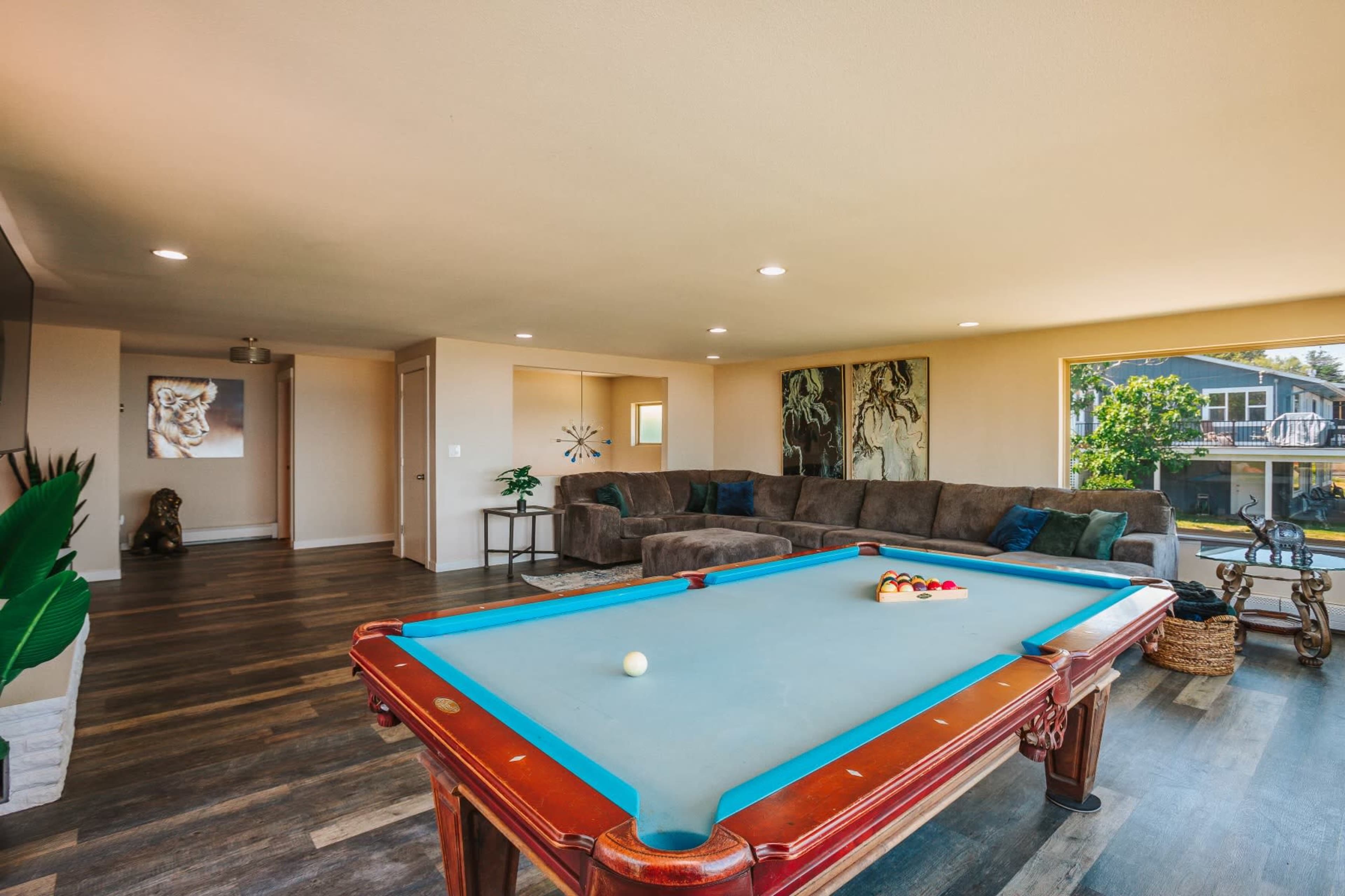 A well-lit room features a billiard table in the foreground and a large sectional couch against the wall, with artwork and decorative elements around.