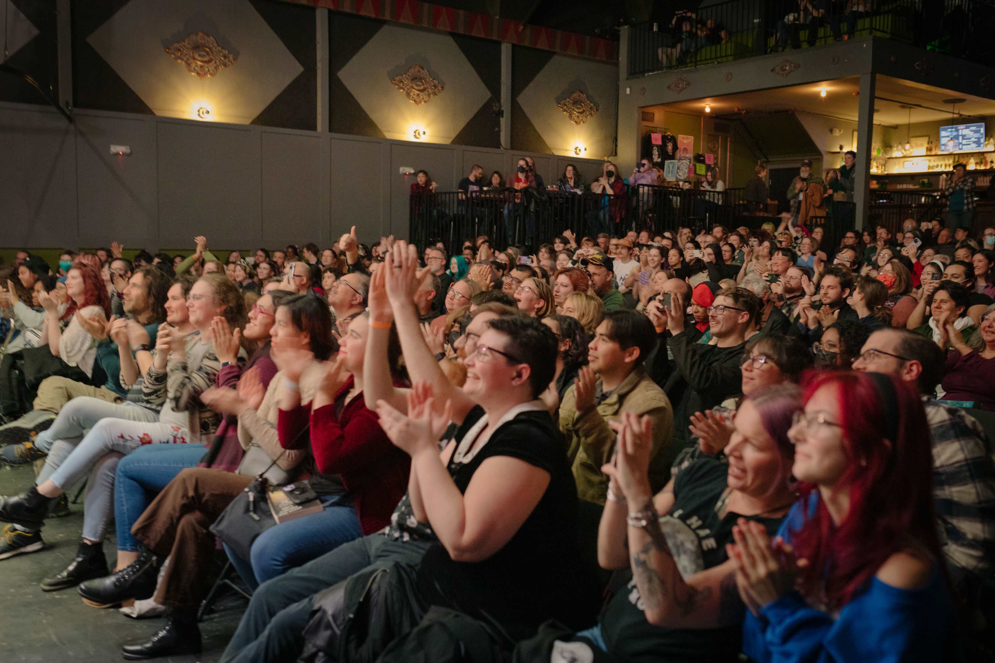 A large audience is clapping and enjoying a performance in a theater setting.
