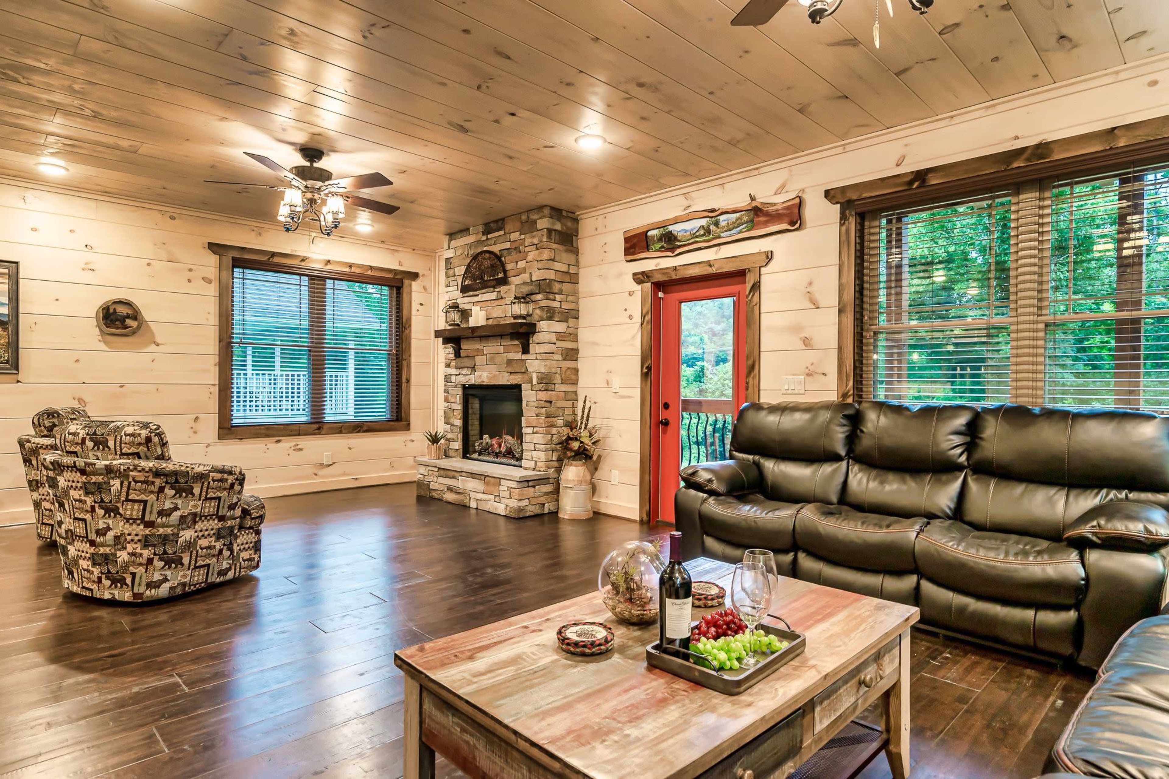 A cozy living room features wooden walls, a stone fireplace, leather seating, and a coffee table with snacks and drinks.
