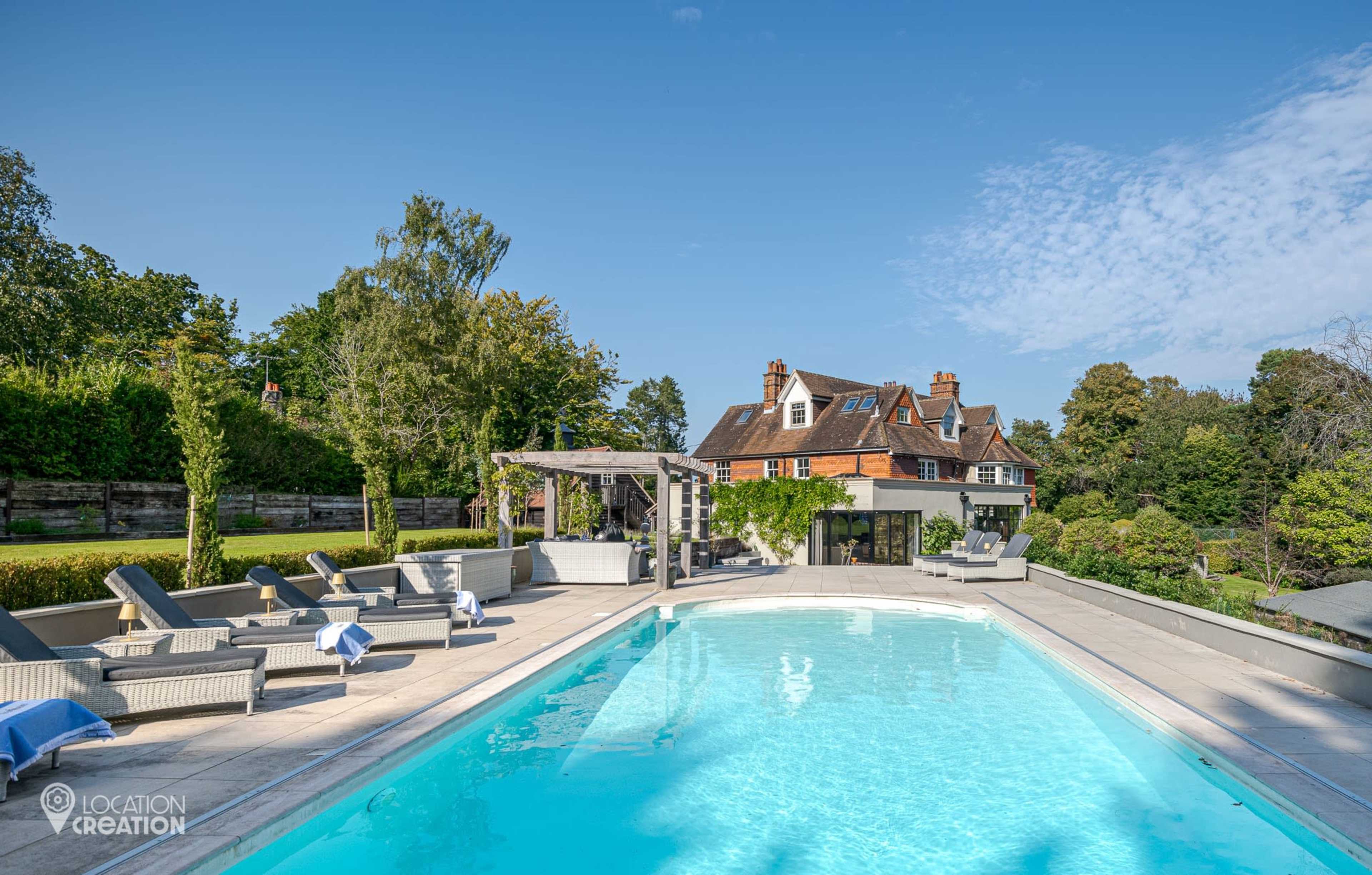The image shows a modern swimming pool surrounded by lounge chairs and a landscaped garden, with a large house visible in the background.