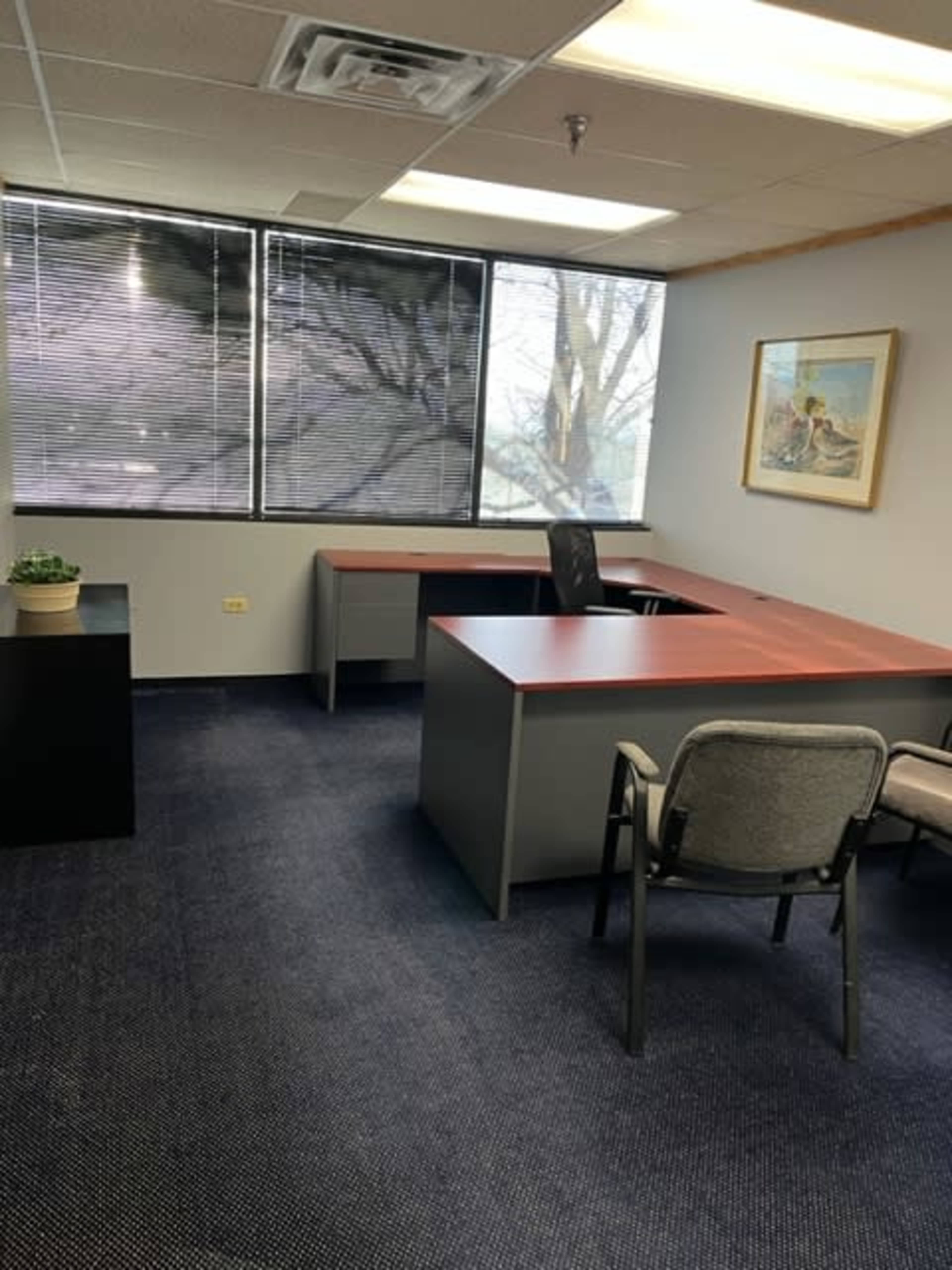 The image shows an empty office space with two desks, a chair, and large windows allowing natural light, framed by blinds.