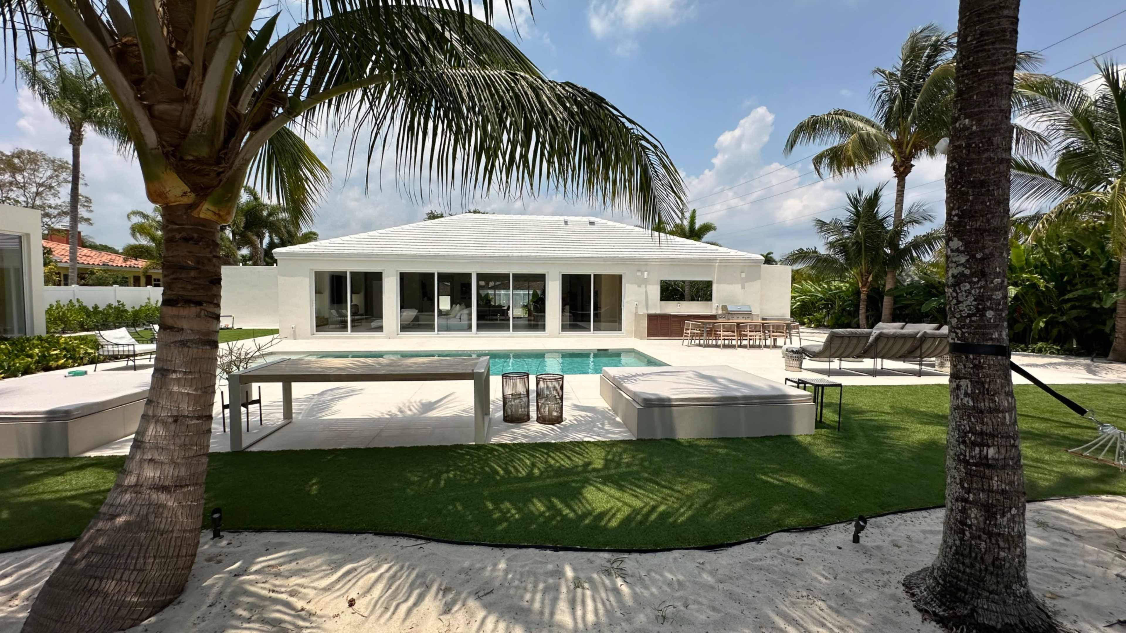 The image shows a modern house with large glass windows, a swimming pool, and a landscaped outdoor area featuring palm trees and lounge furniture.