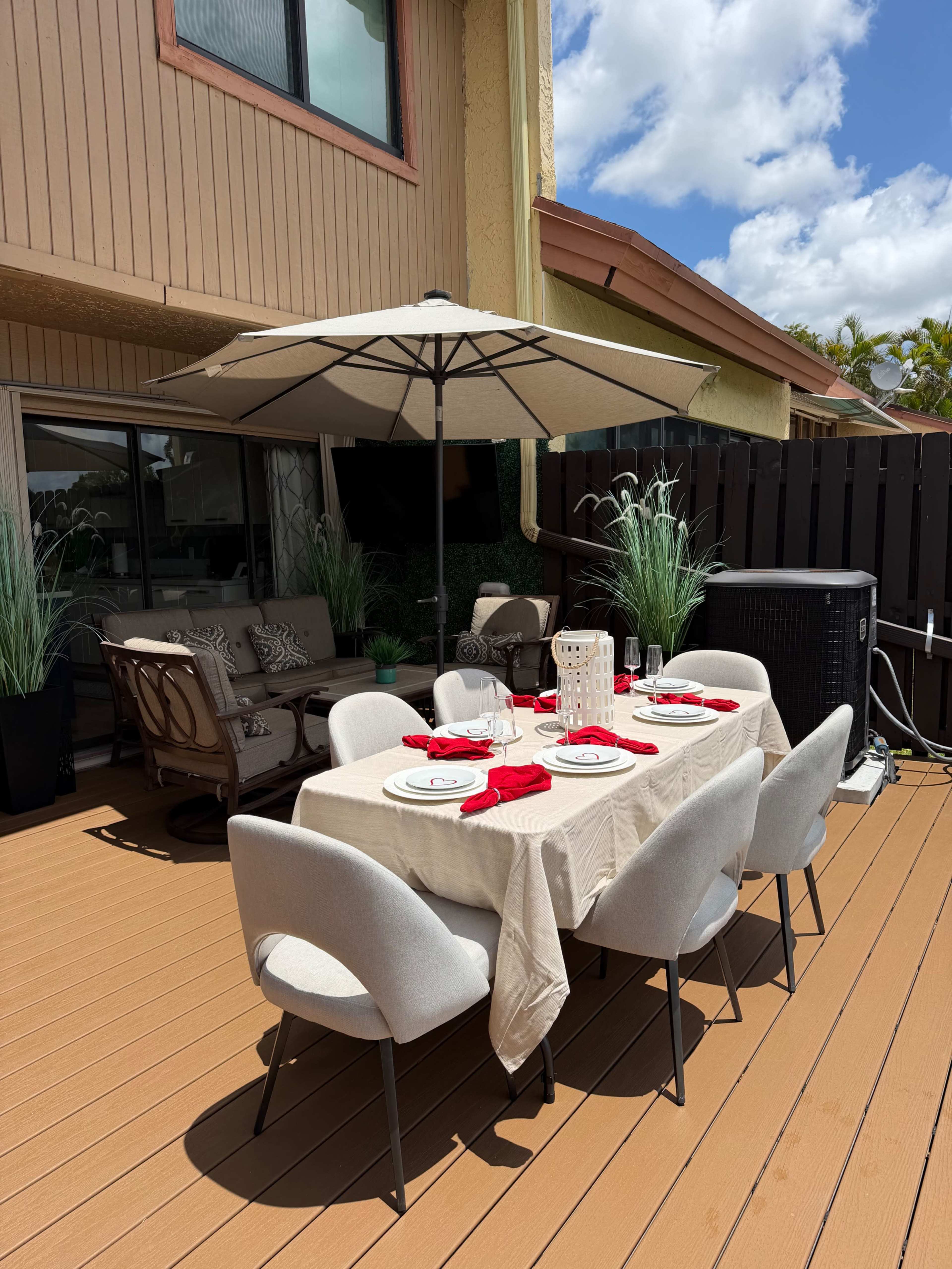 A dining table set with red napkins and dinnerware is arranged under a large umbrella on a patio with potted plants and outdoor furniture.