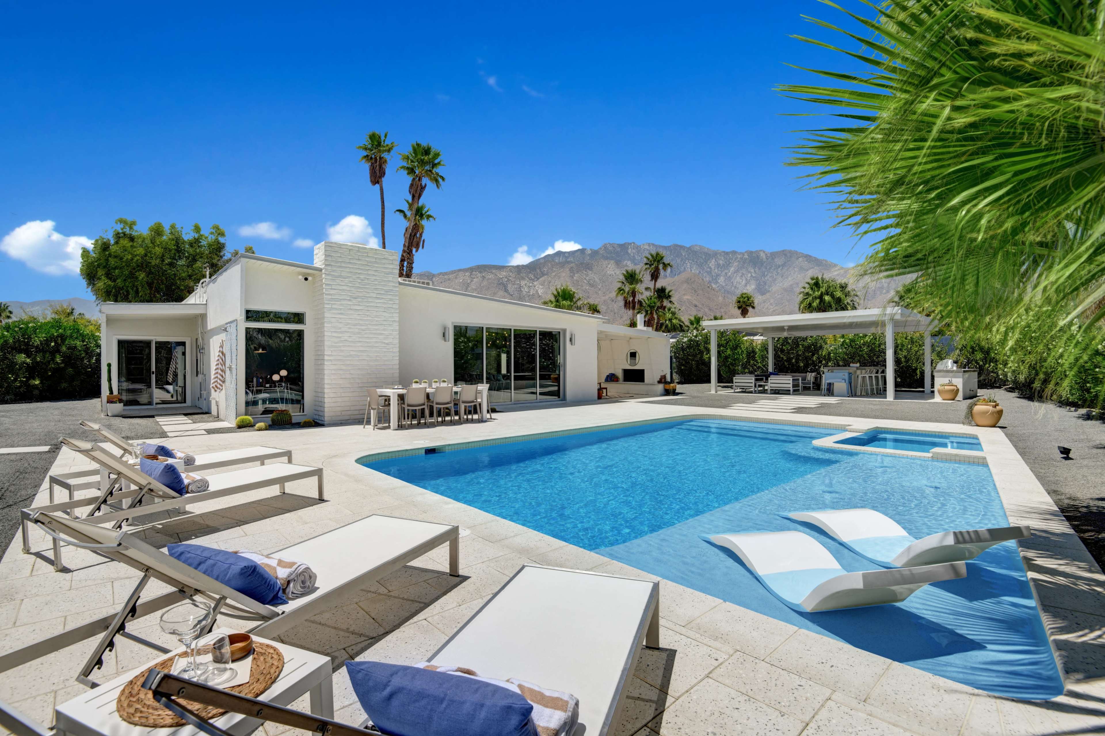 A modern white villa with a swimming pool and lounge chairs is surrounded by palm trees and mountains under a clear blue sky.