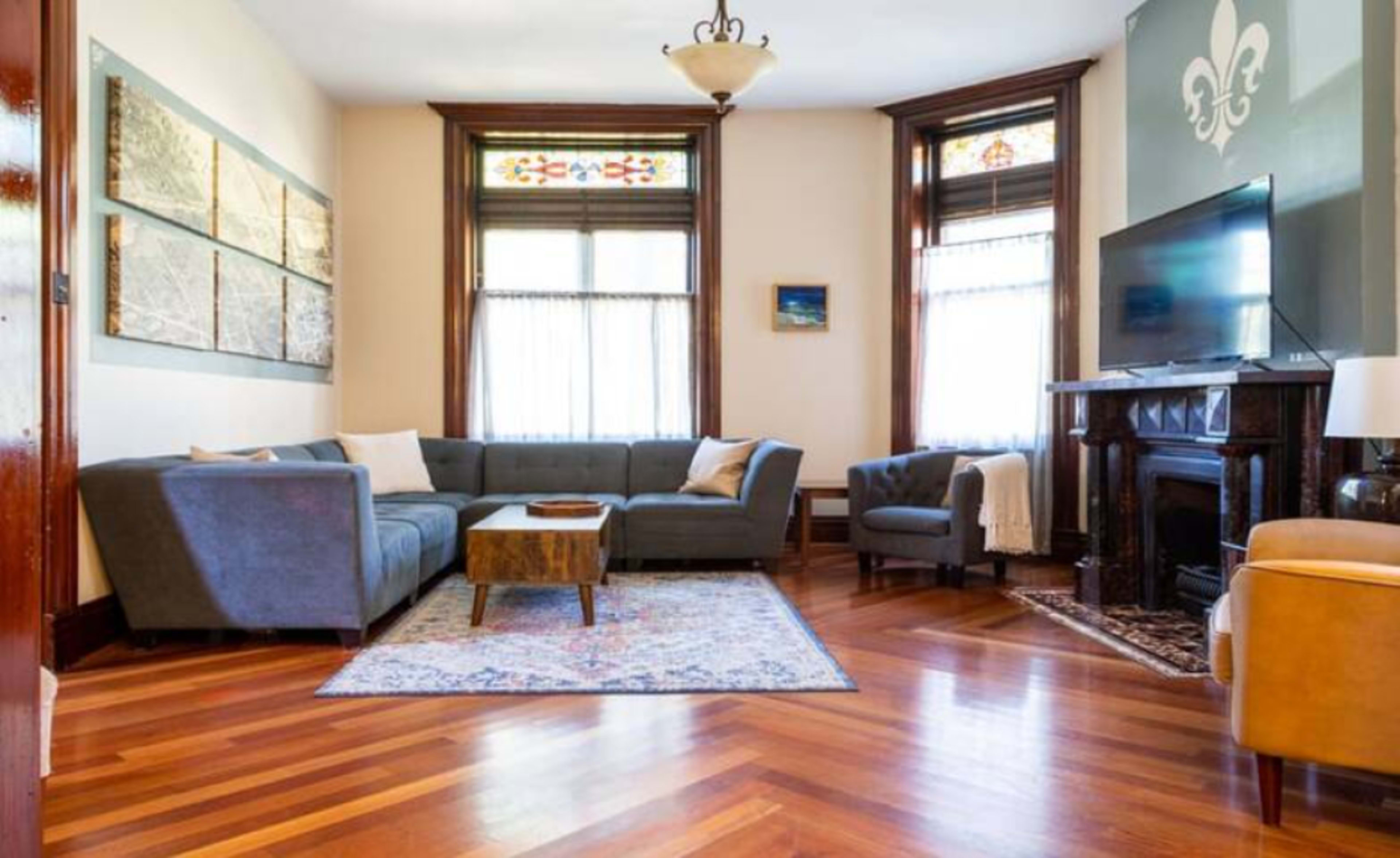 The image depicts a living room featuring a large gray sectional sofa, a wooden coffee table, a television, and a decorative fireplace, with hardwood floors and large windows allowing natural light.
