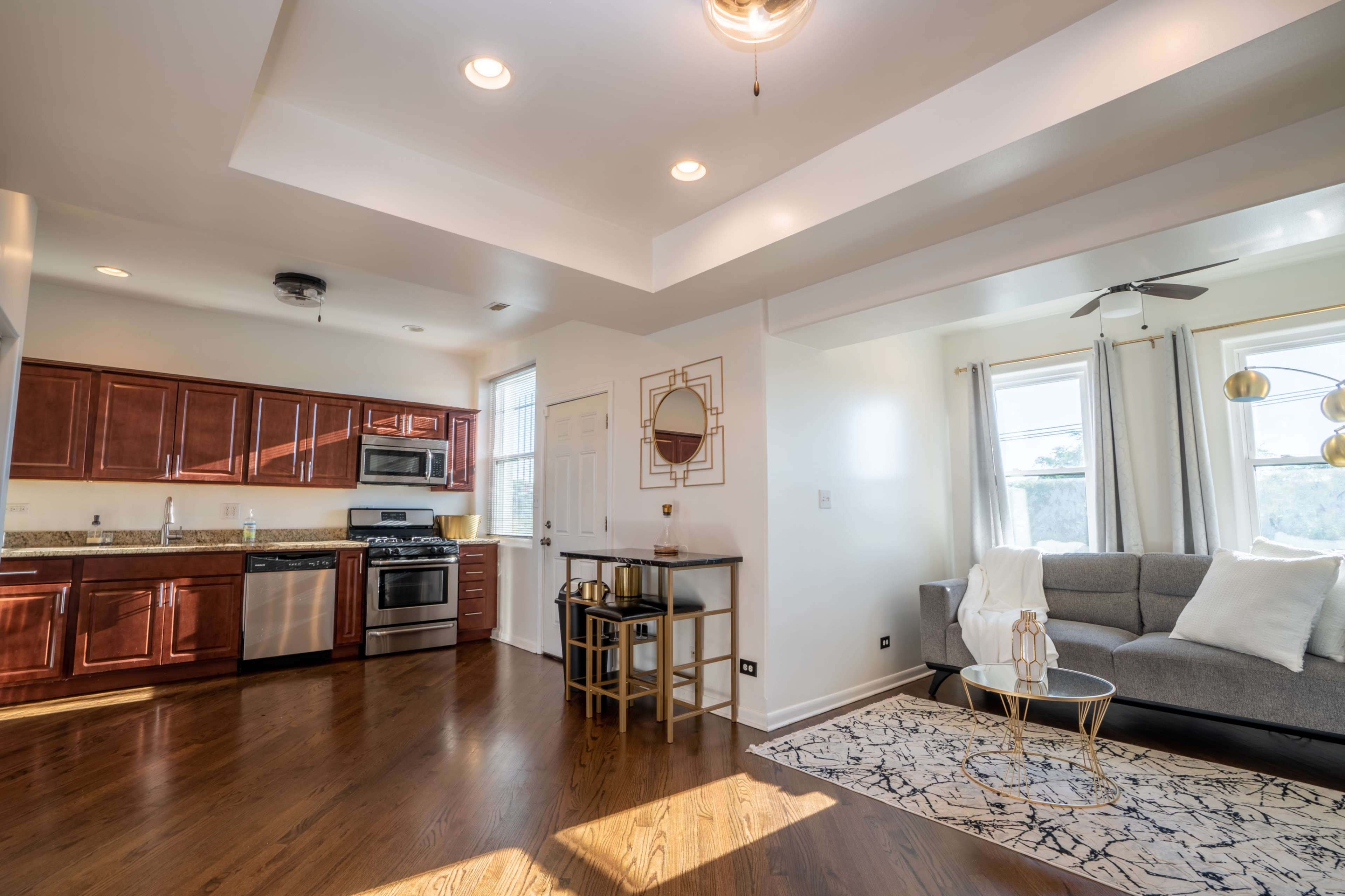 A modern kitchen and living area with wooden cabinetry, stainless steel appliances, and a gray sofa facing a coffee table on a patterned rug.