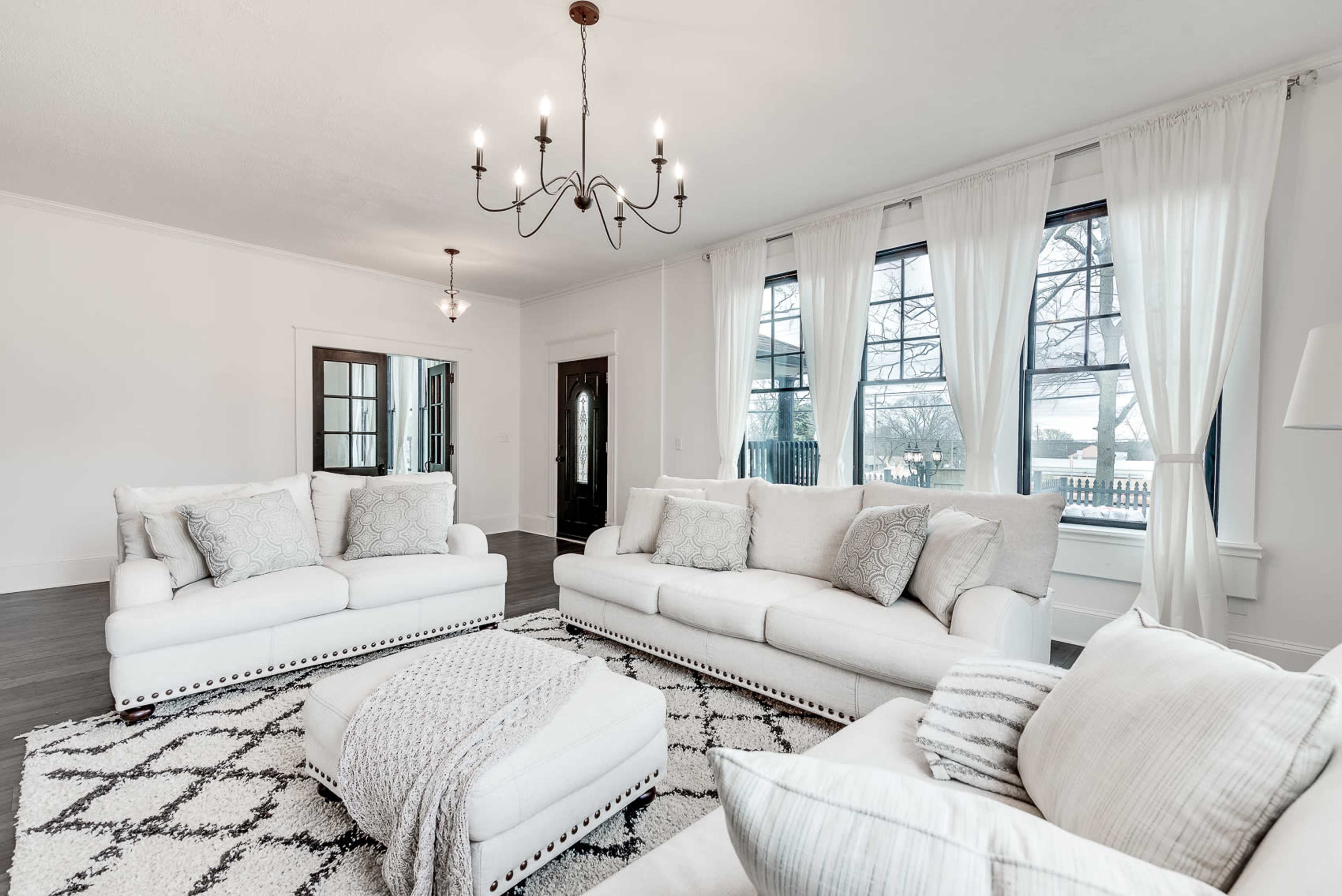 The living room features white sofas, a patterned area rug, and large windows that allow natural light to fill the space.