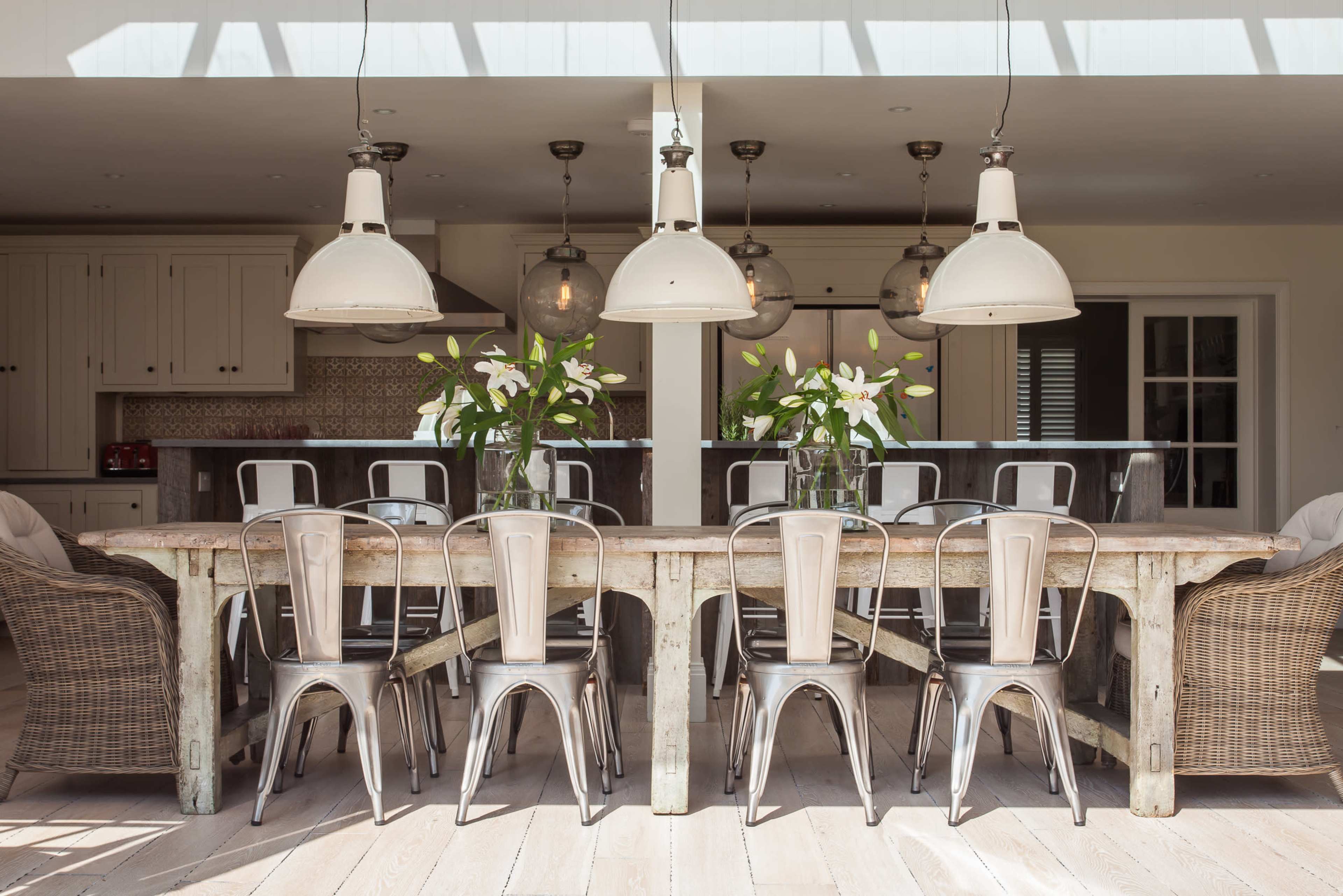 A large wooden dining table surrounded by metal chairs is illuminated by pendant lights, with decorative flowers in vases on the table.