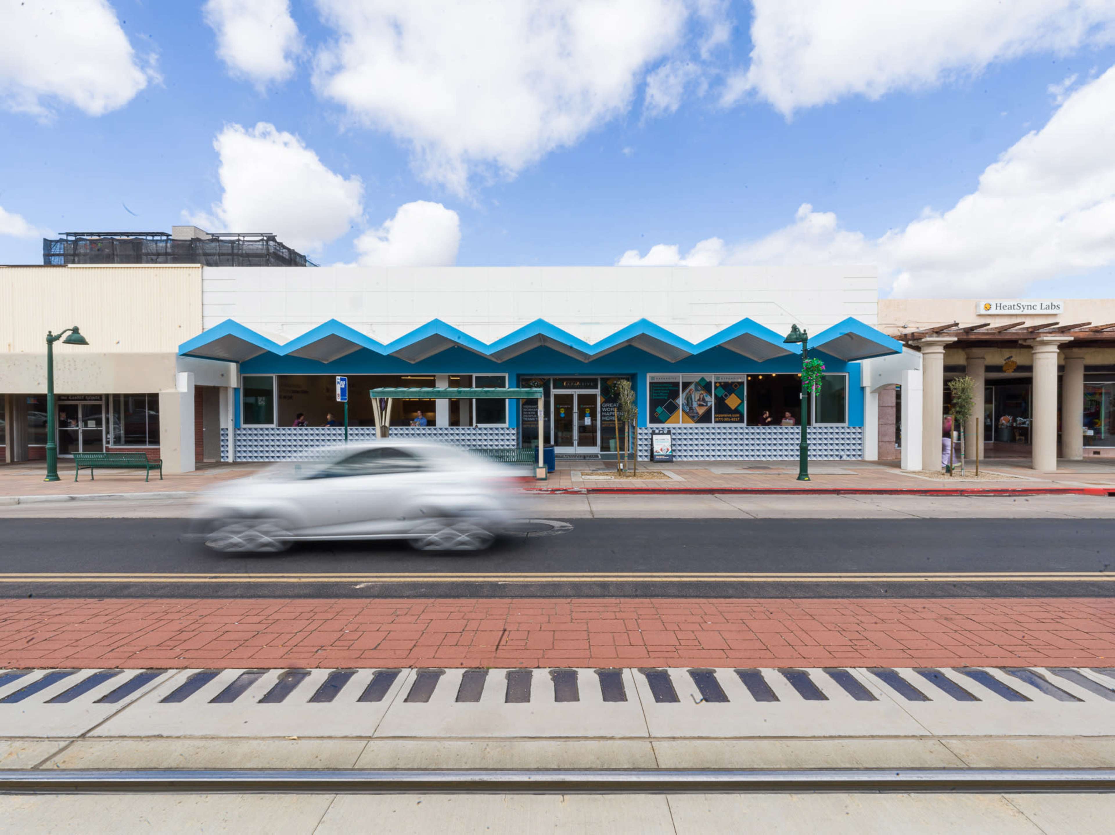 A white car drives past a storefront with a distinctive blue zigzag roof design on a sunny day.