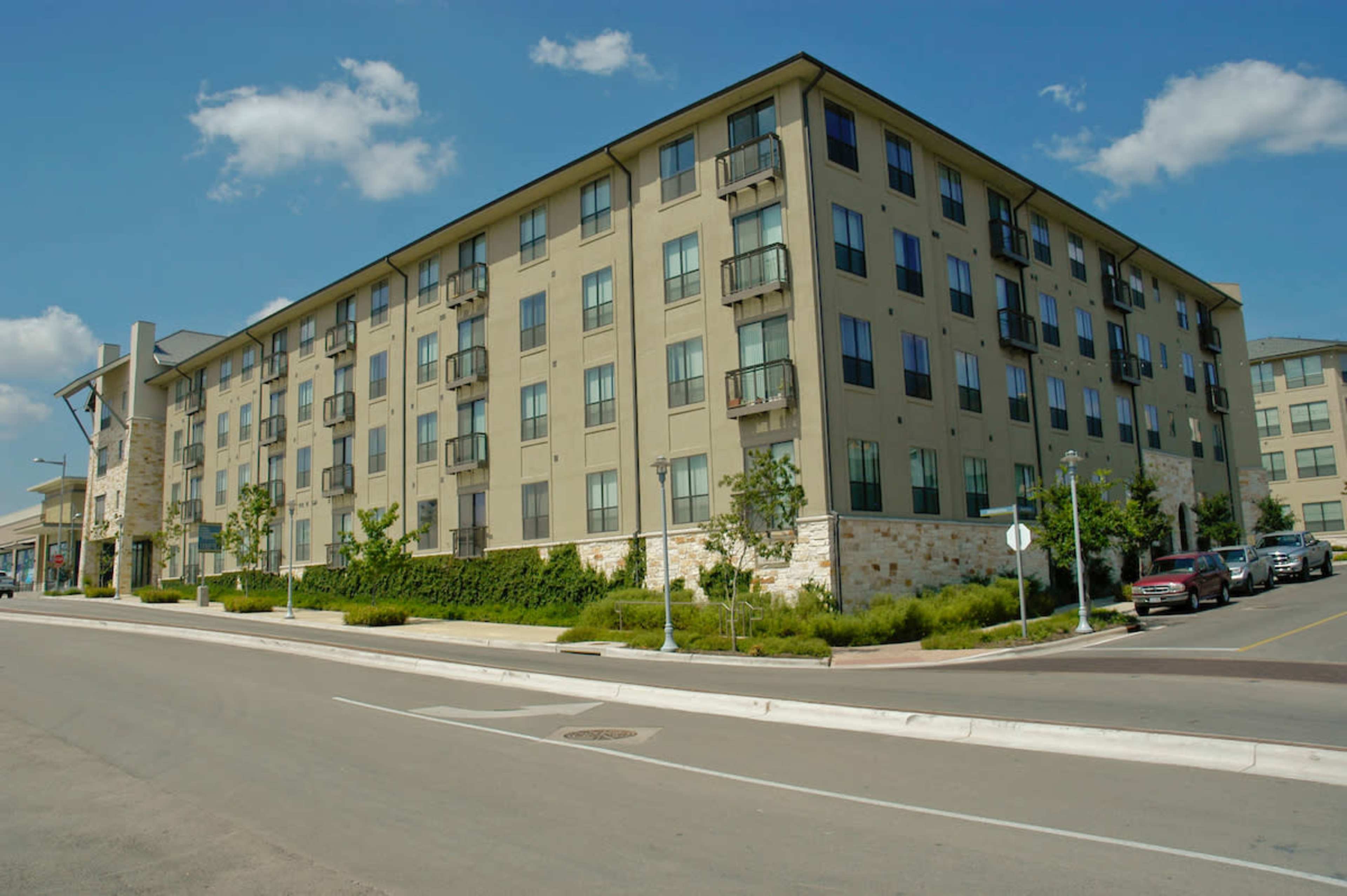 A modern, four-story apartment building with balconies, situated along a paved road with green landscaping and parked cars nearby.