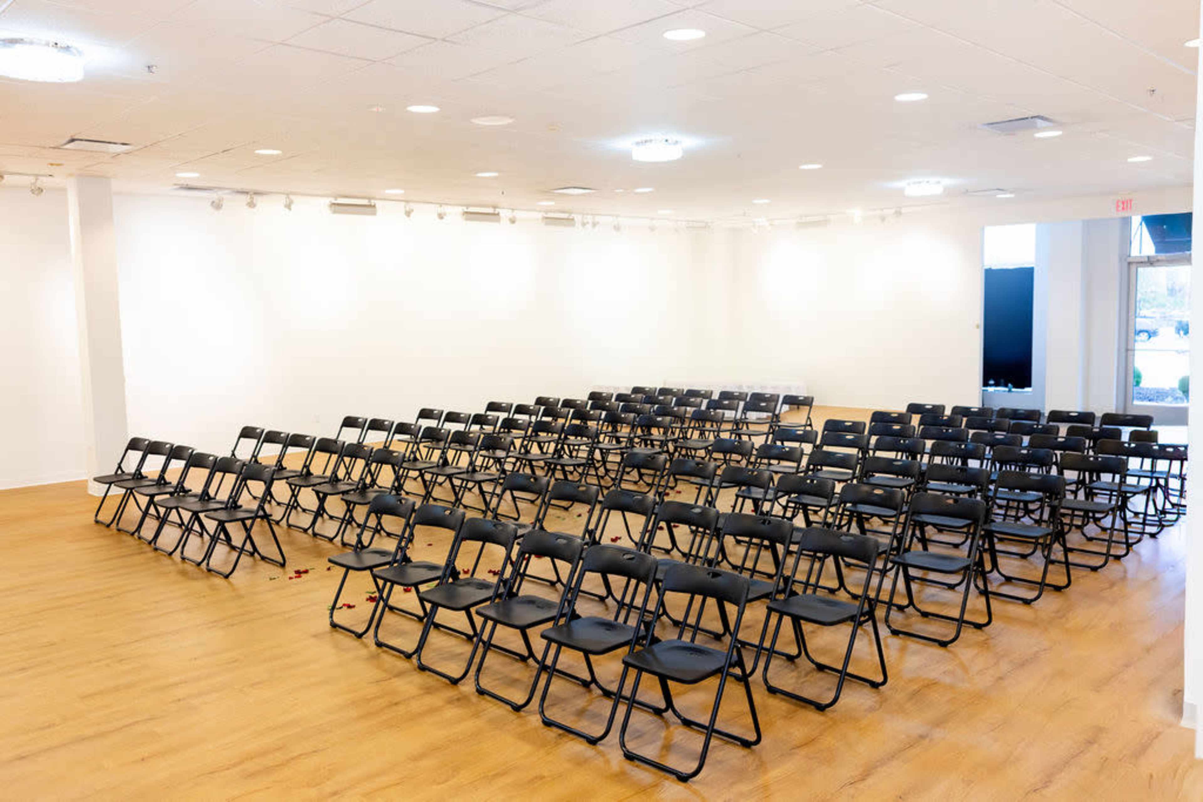 A large room features rows of black folding chairs arranged in a semi-circle facing a blank wall.