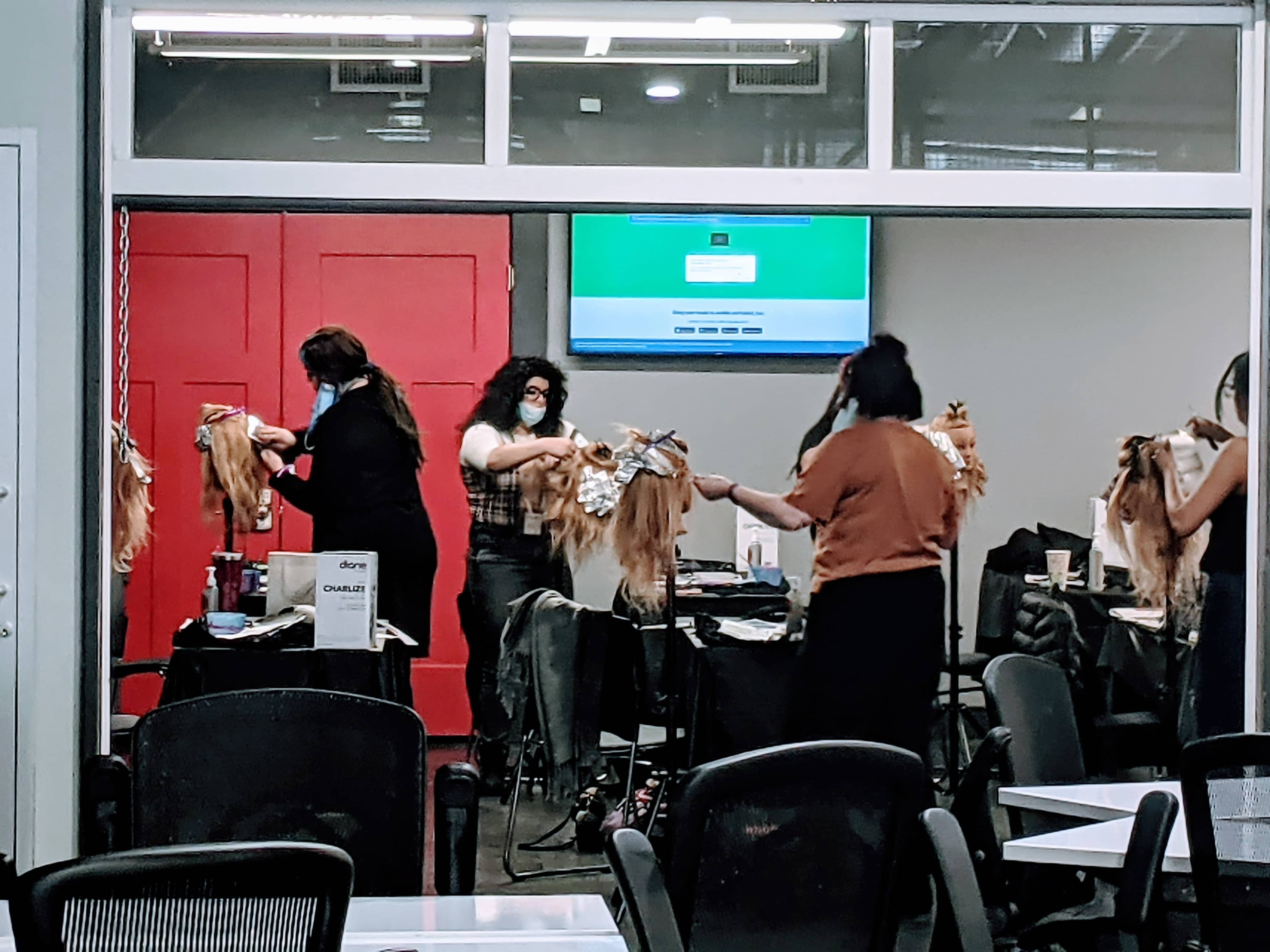 A group of hairstylists in an indoor salon setting are working on mannequins with long hair, using various styling tools.