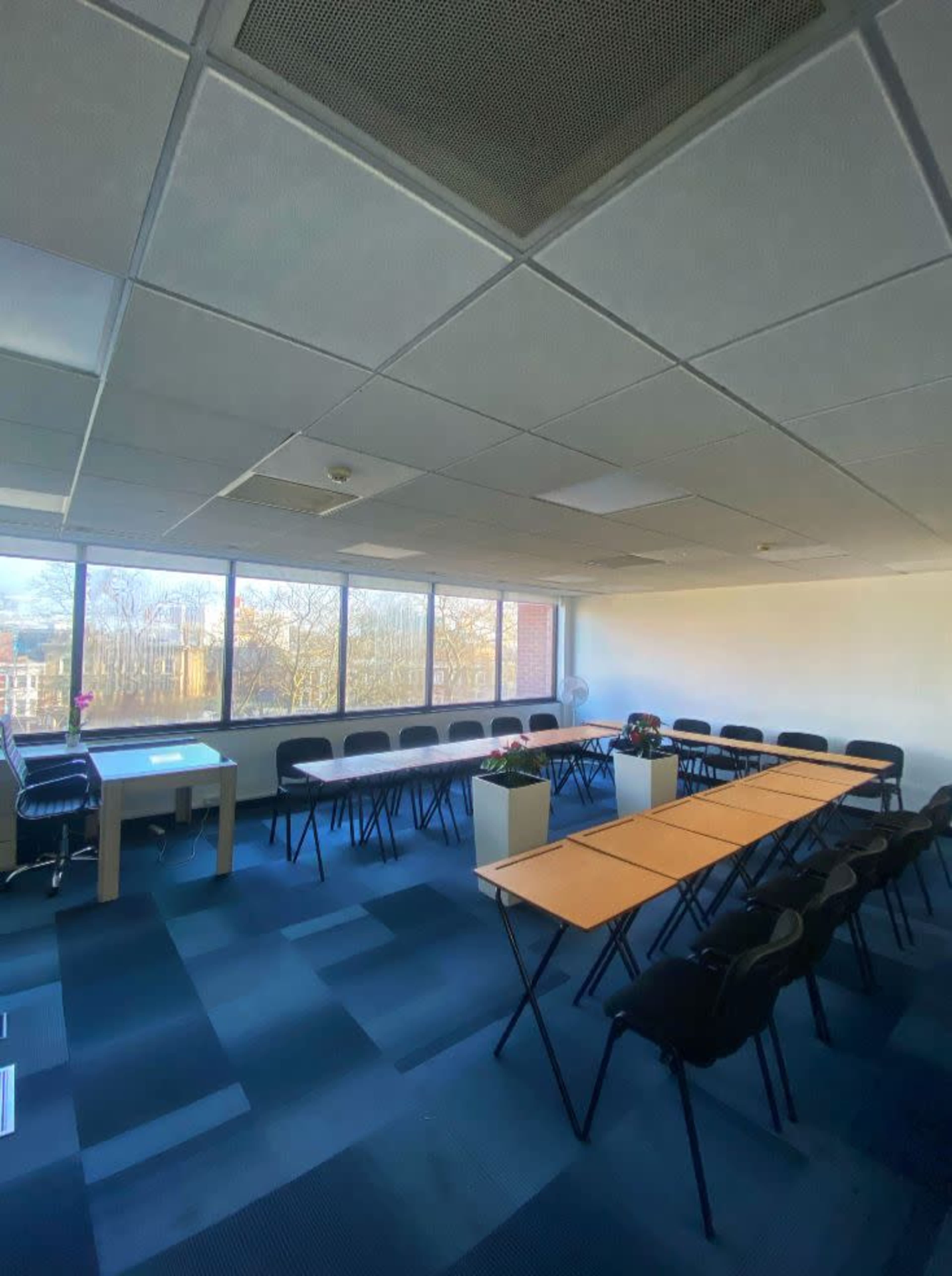 The image shows a conference room with a long table set up in the center, surrounded by chairs, and large windows letting in natural light.