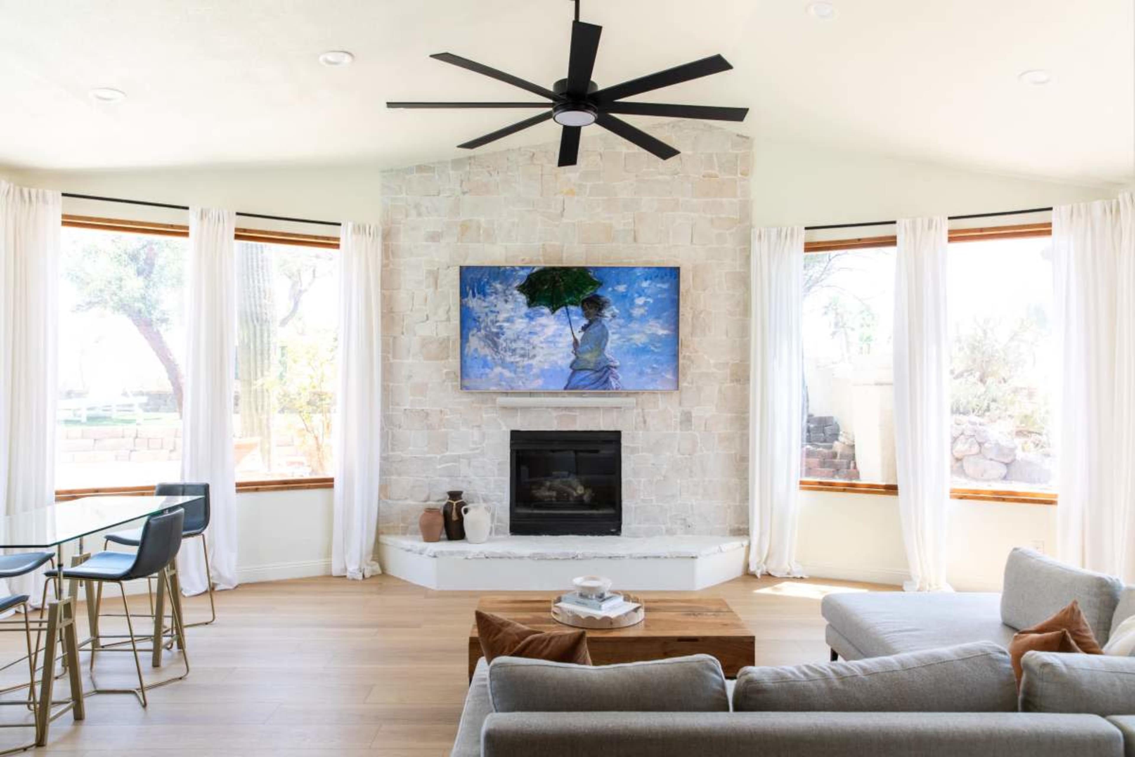 A modern living room features a stone fireplace with a mounted TV above it, large windows with sheer curtains, and a comfortable gray sofa.