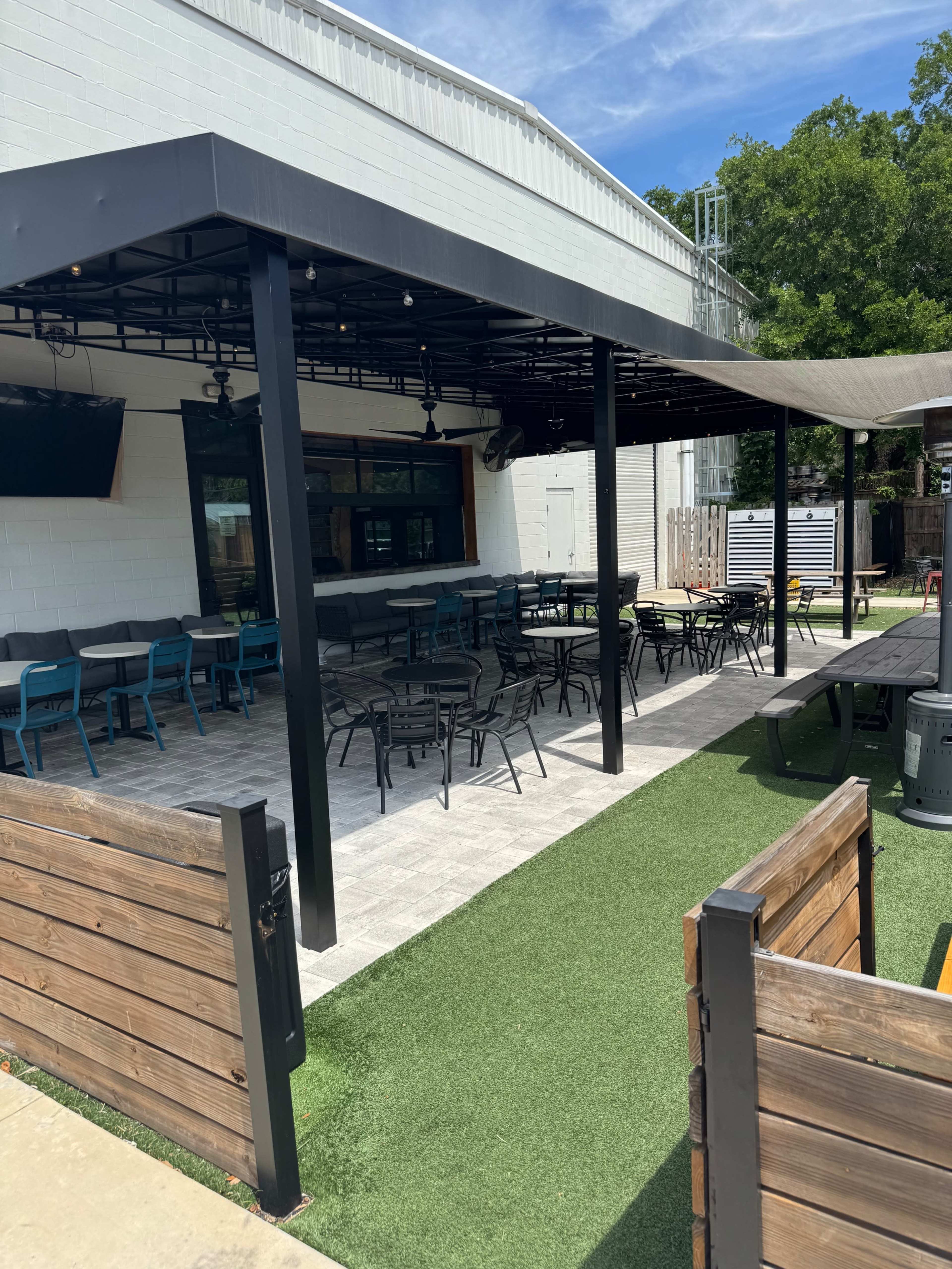 The image shows a spacious outdoor seating area with metal tables and chairs under a canopy, adjacent to a building with a large window.