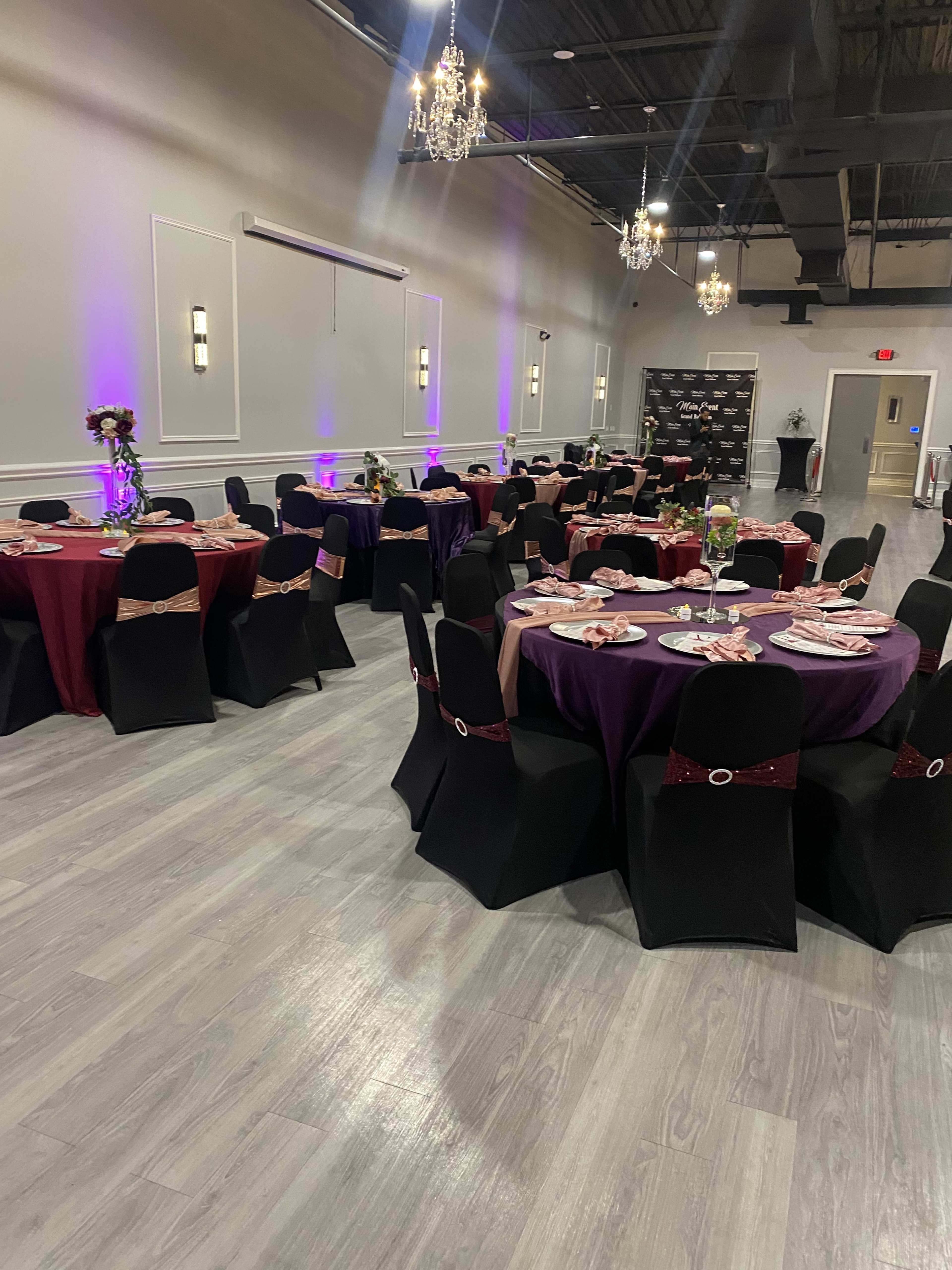 A banquet hall set up with round tables covered in black and burgundy tablecloths, adorned with centerpieces and place settings, under decorative chandeliers.