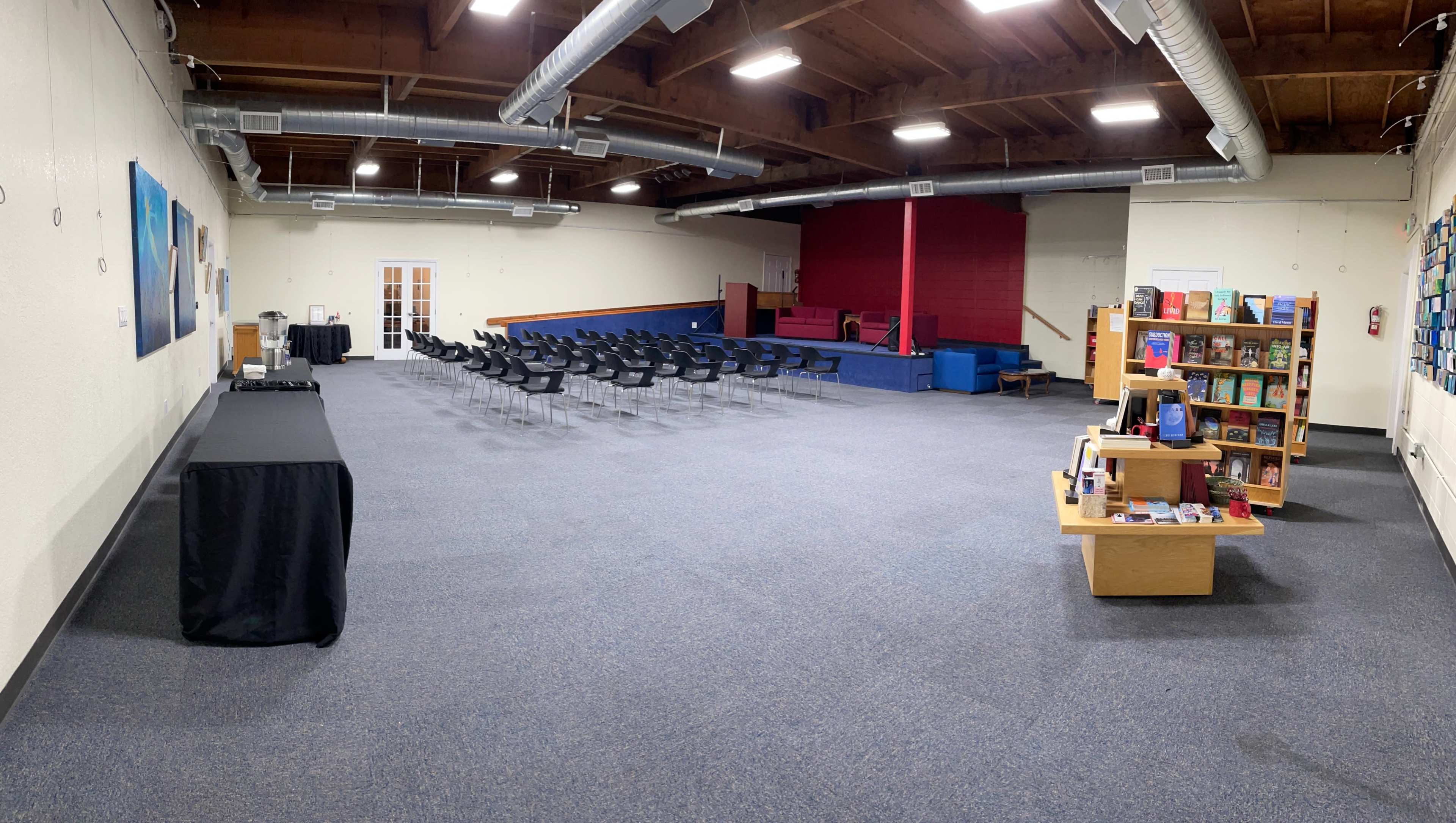 A spacious room with a row of black chairs facing a stage, along with a table and a bookshelf filled with books along the side.