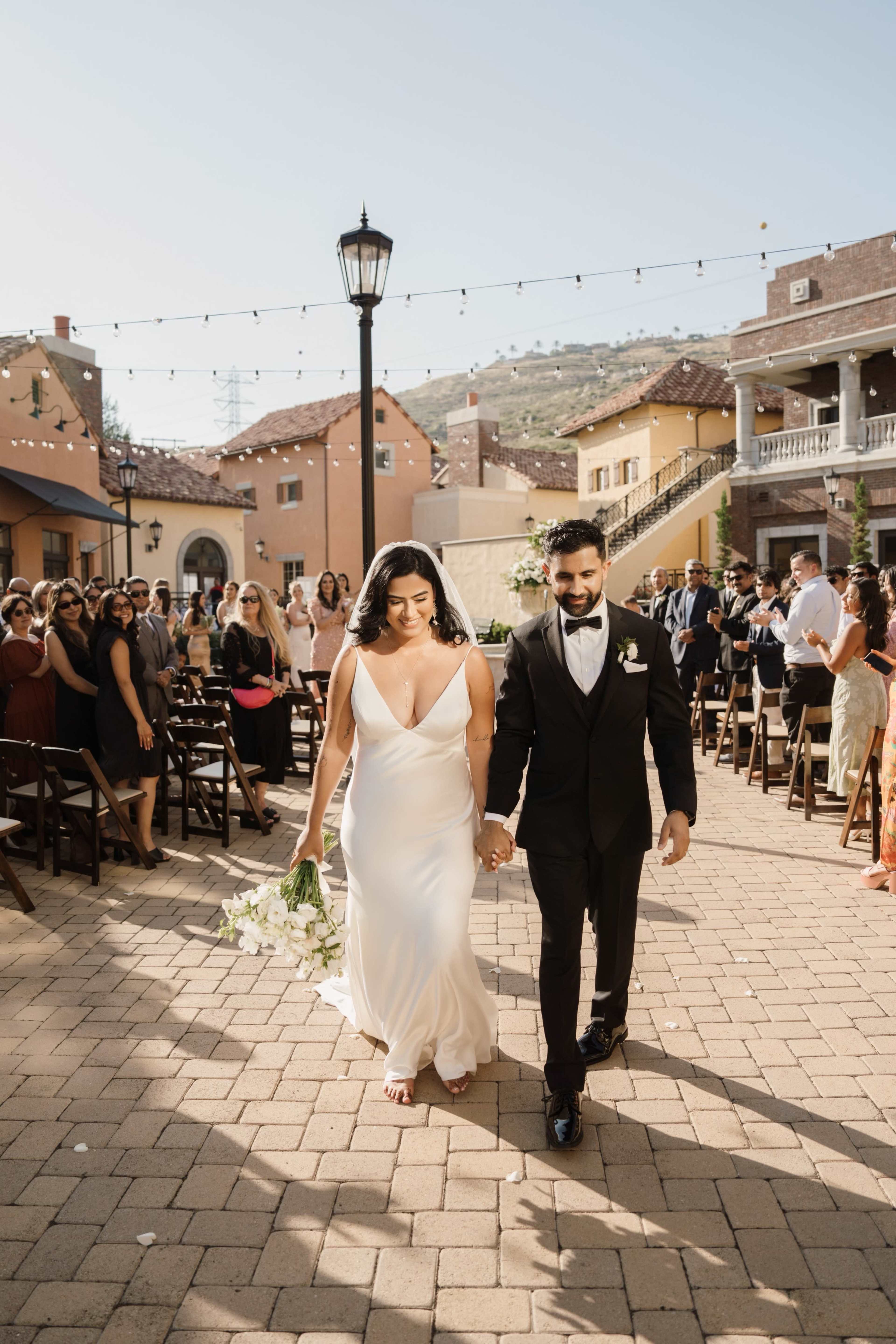 A newly married couple walks hand-in-hand down the aisle, surrounded by seated guests at an outdoor wedding venue.