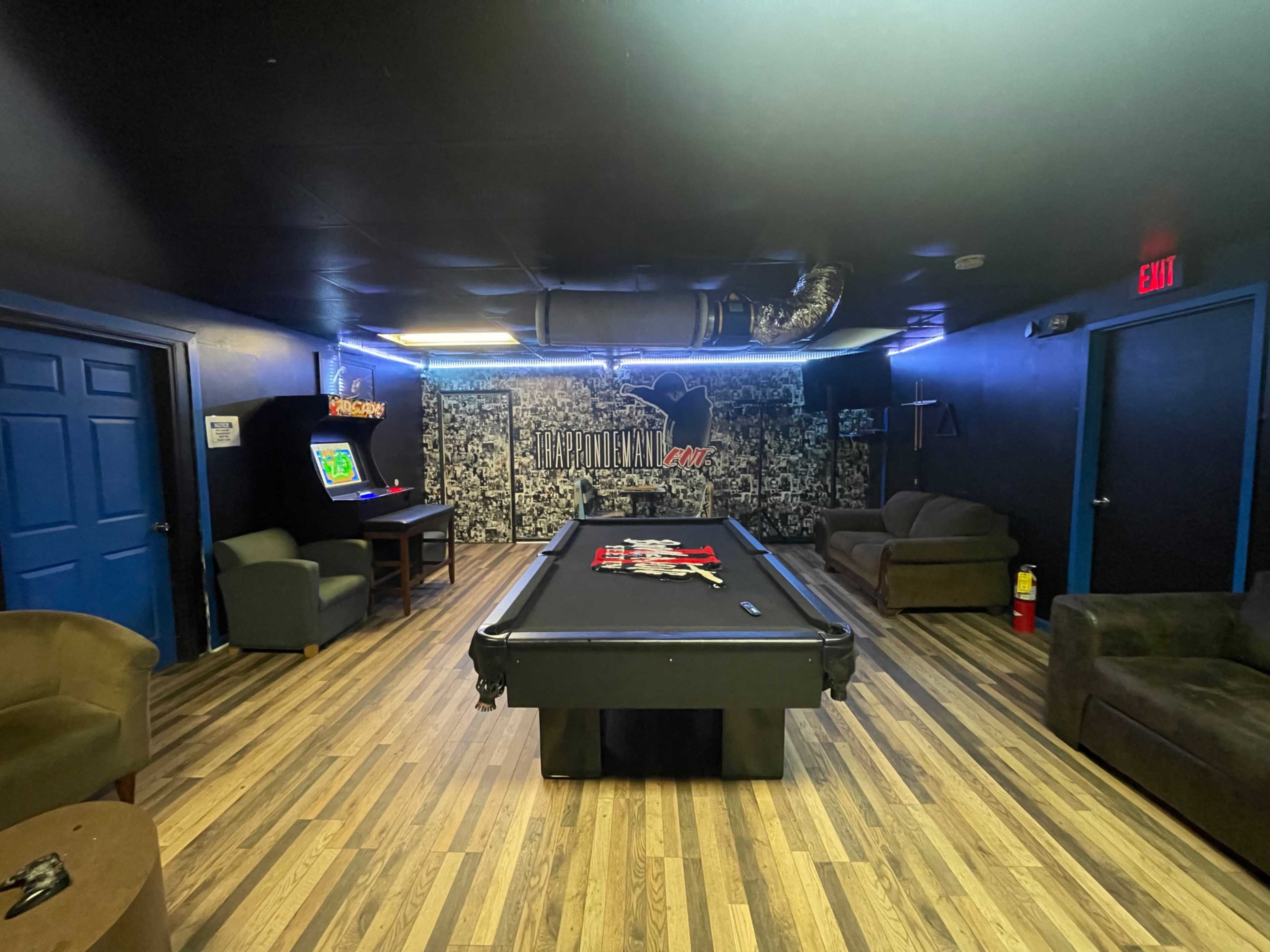 A game room featuring a pool table in the center, surrounded by two armchairs, a couch, and an arcade machine in the corner, with blue walls and wooden flooring.