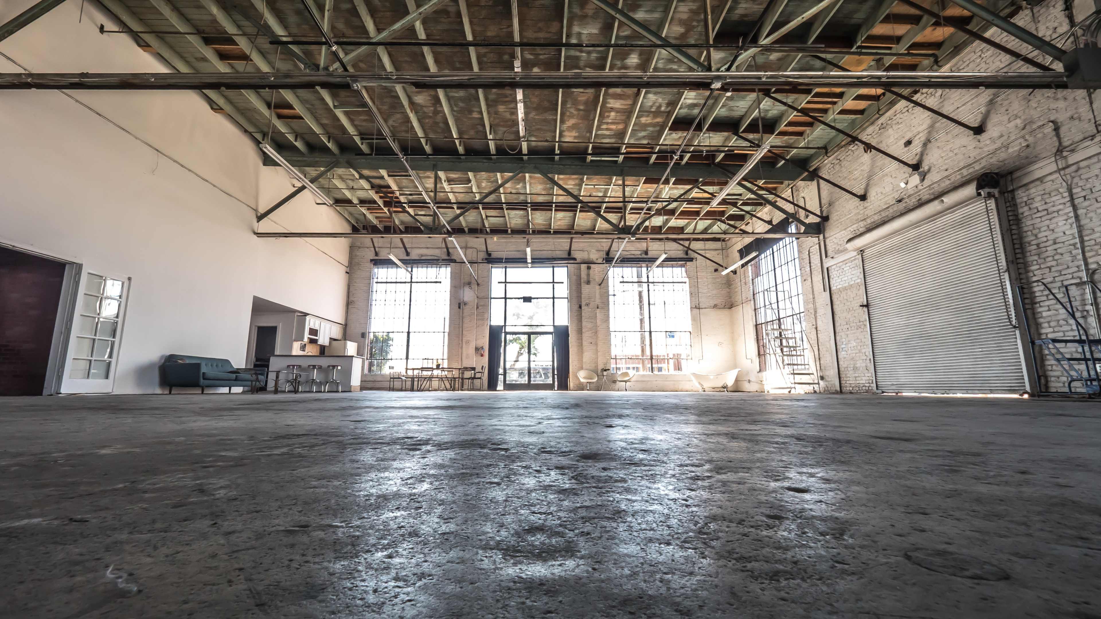 The image shows a spacious, empty industrial interior with large windows, concrete flooring, and an exposed ceiling.
