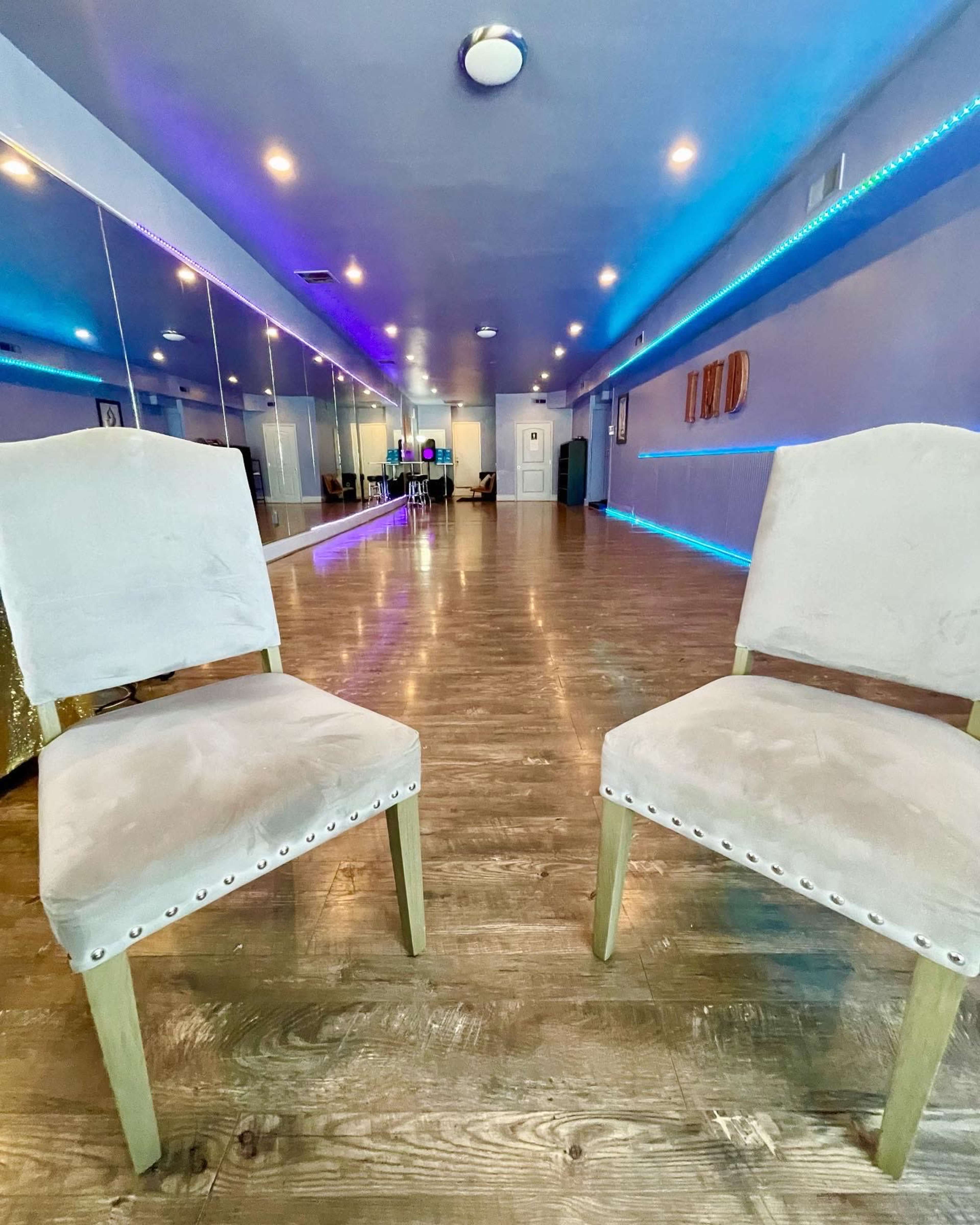 Two upholstered chairs with decorative nails are positioned facing each other in a brightly lit room featuring mirrored walls and colorful LED lights.