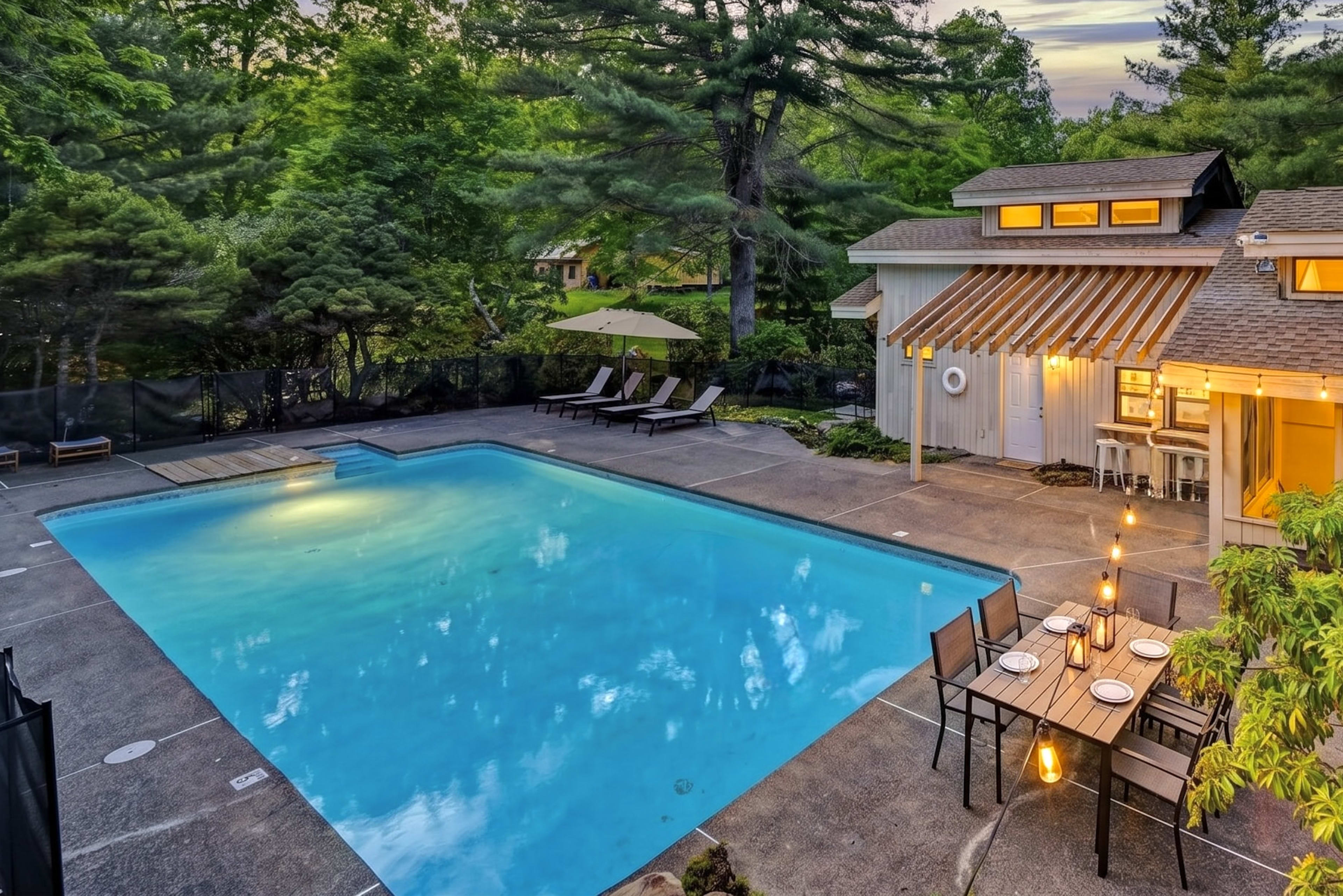 The image shows a spacious outdoor area featuring a swimming pool surrounded by lounge chairs and a dining table, with a house in the background and trees lining the property.